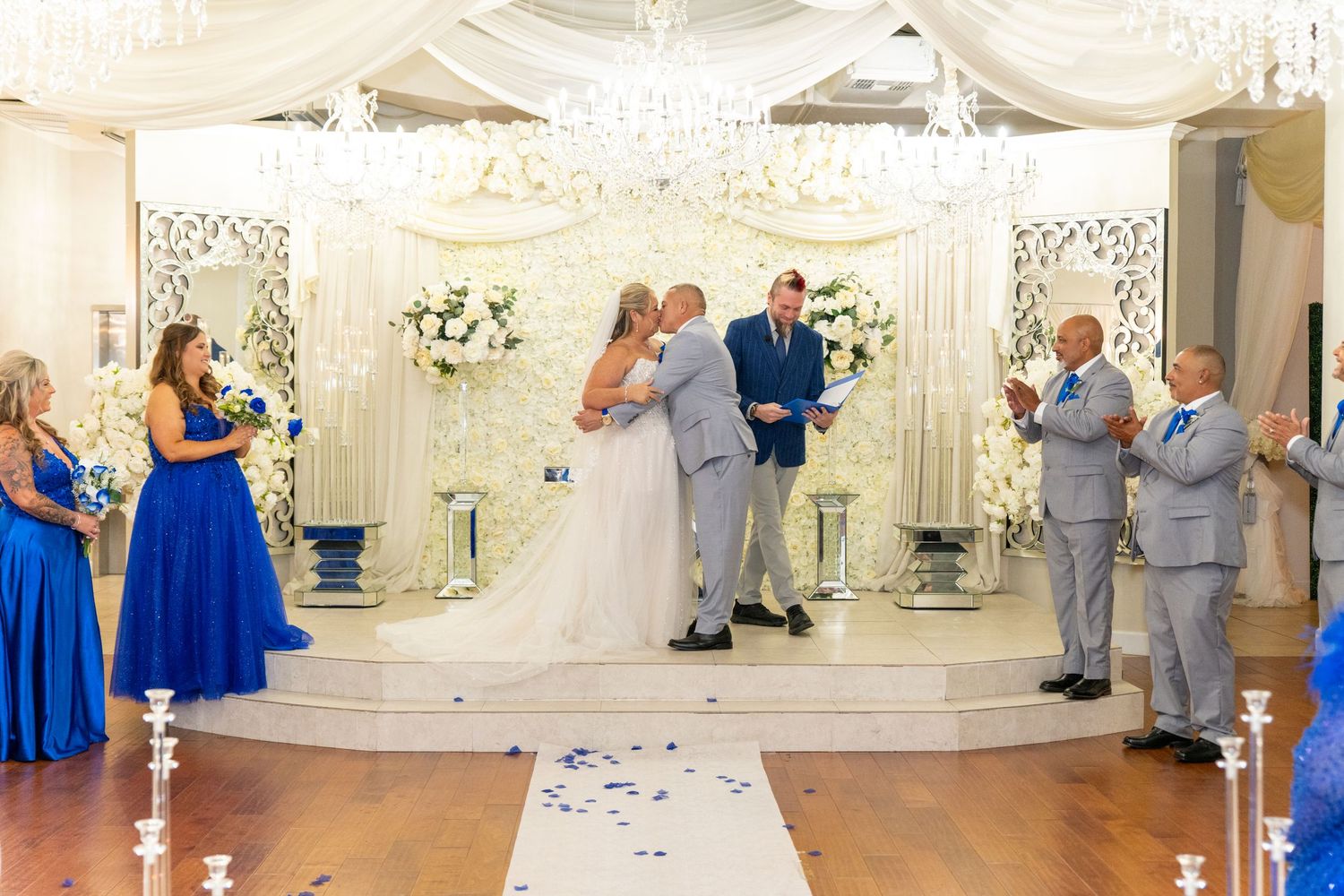 Bride and groom share first kiss at elegant wedding ceremony with blue bridesmaids, chandelier, and white floral decor.