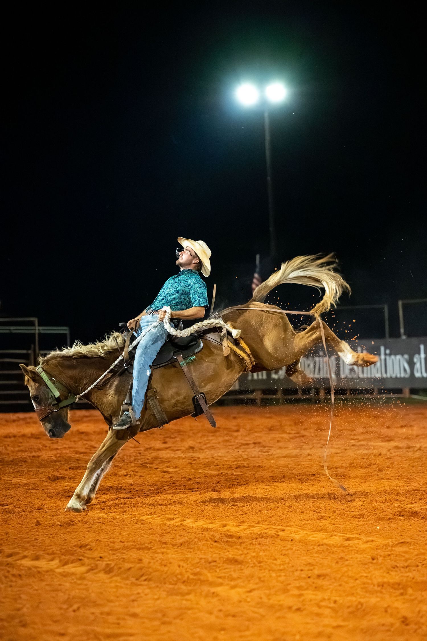 A cowboy in a teal shirt and white hat rides a bucking bronco at a night rodeo on red dirt arena.