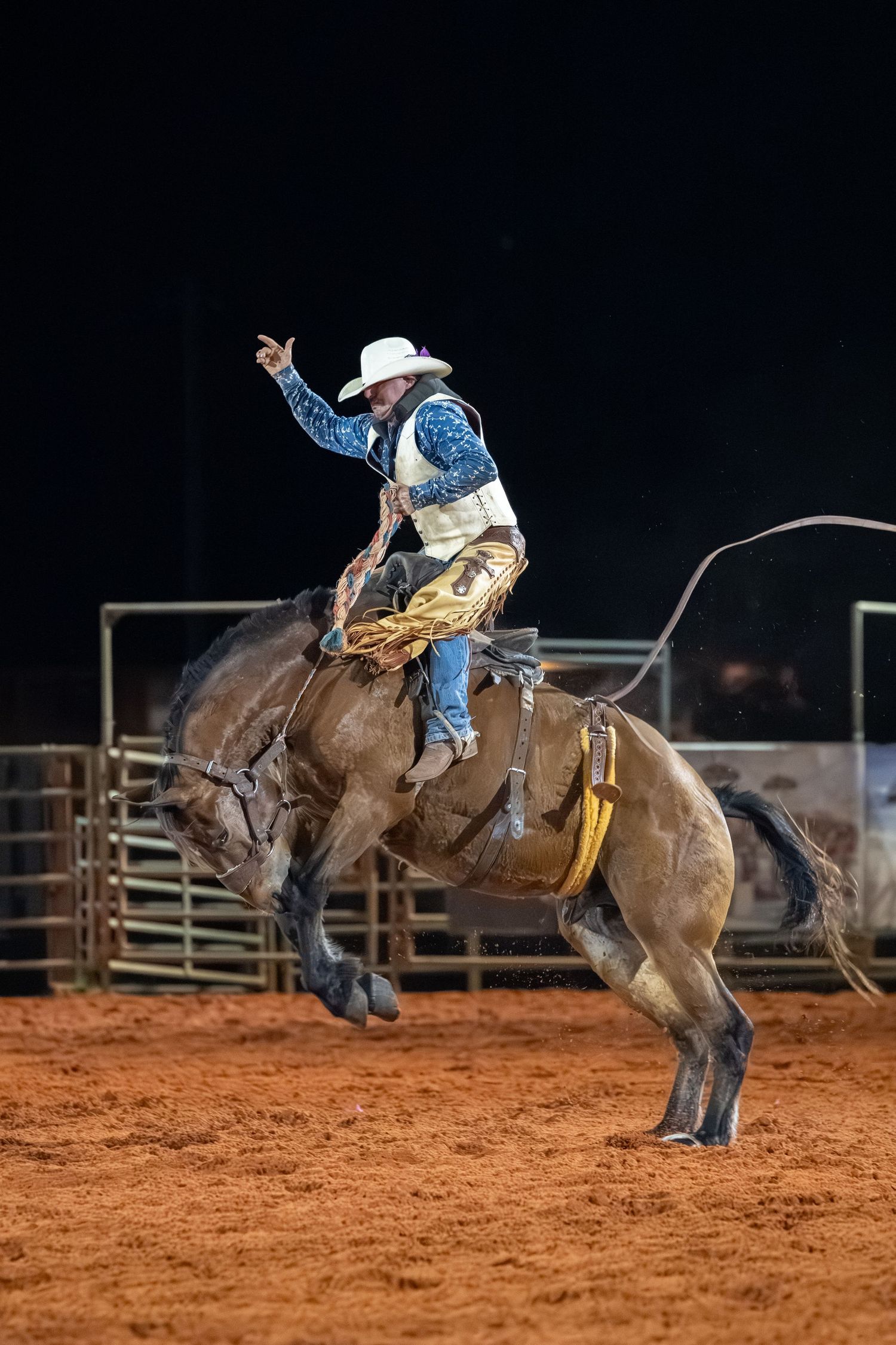Cowboy riding a bucking bronco at a nighttime rodeo competition on red dirt arena.
