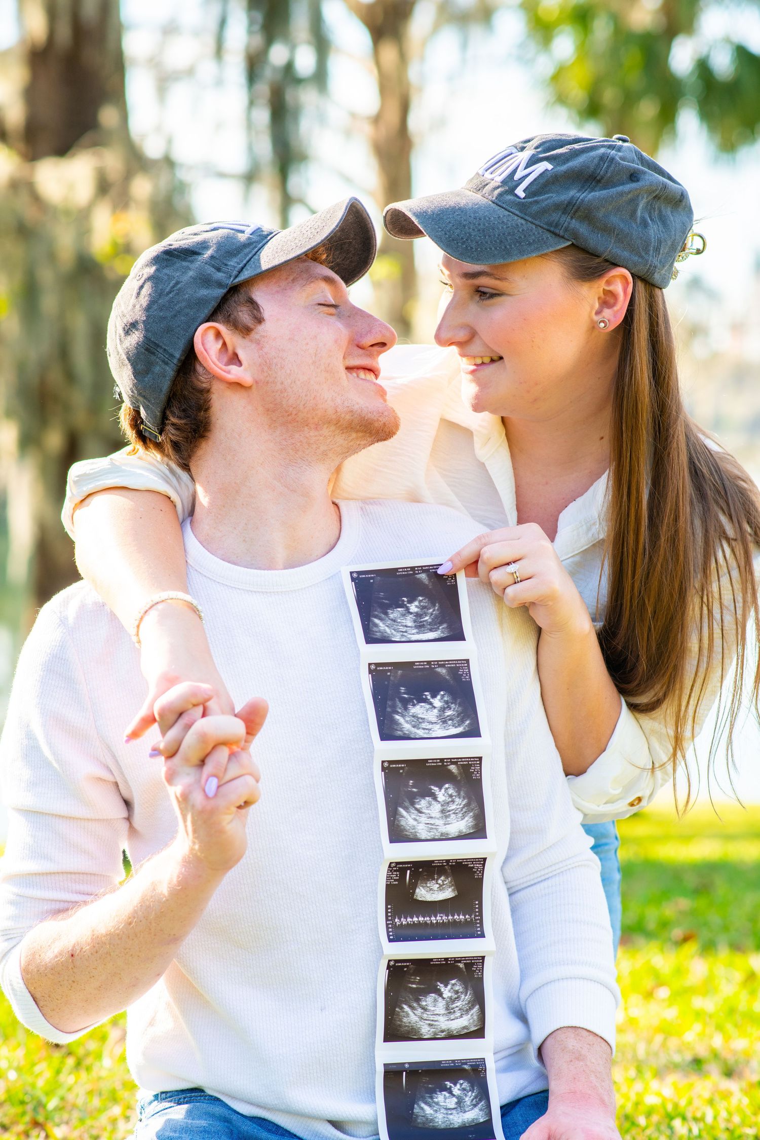 Happy couple holding ultrasound photos outdoors, wearing matching caps, smiling at each other.