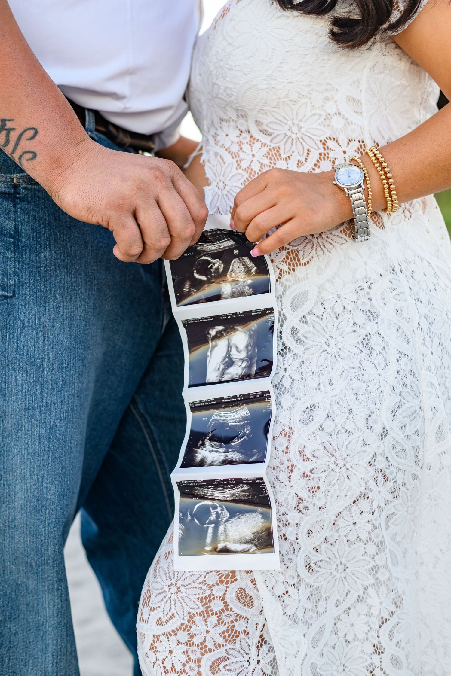 Expecting couple in jeans and a white lace dress holding a strip of ultrasound photos together in a maternity announcement.