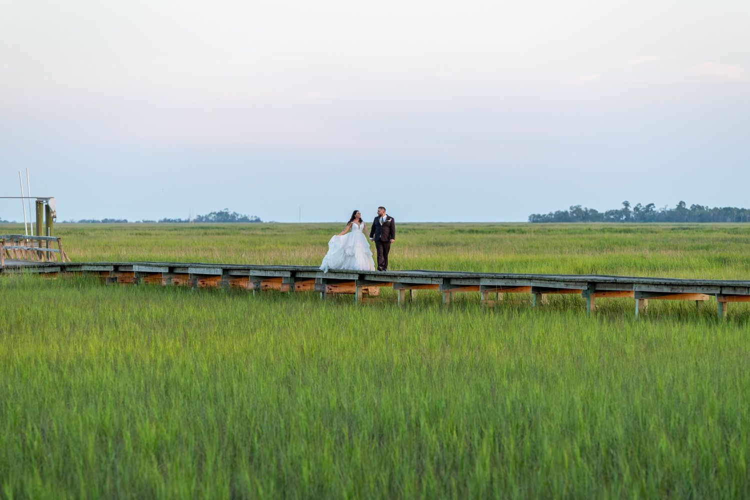 Bride and groom walk along a wooden boardwalk through lush green marshlands during their outdoor wedding ceremony.