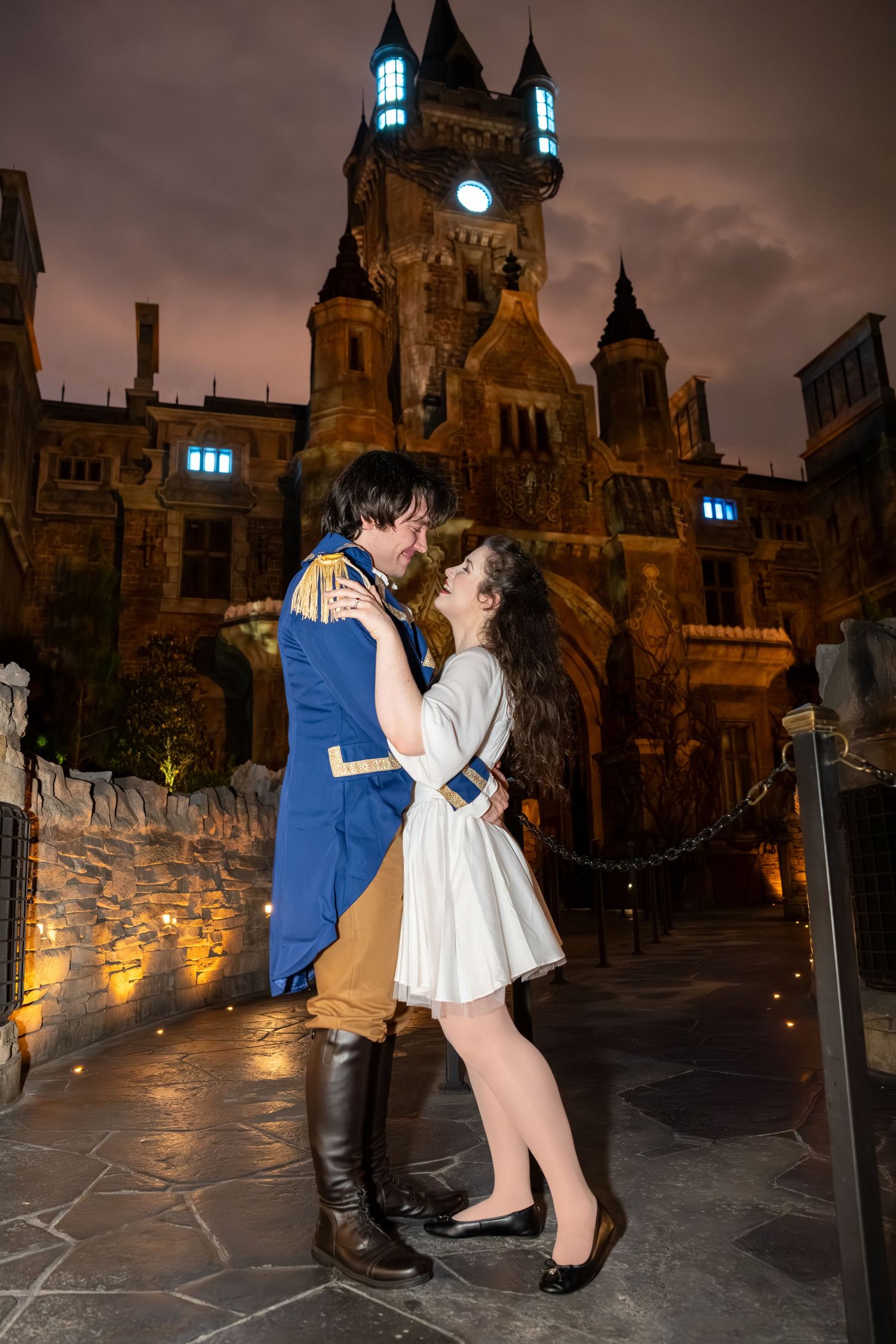 Couple in Beauty and the Beast costumes embrace romantically before an illuminated castle at night.