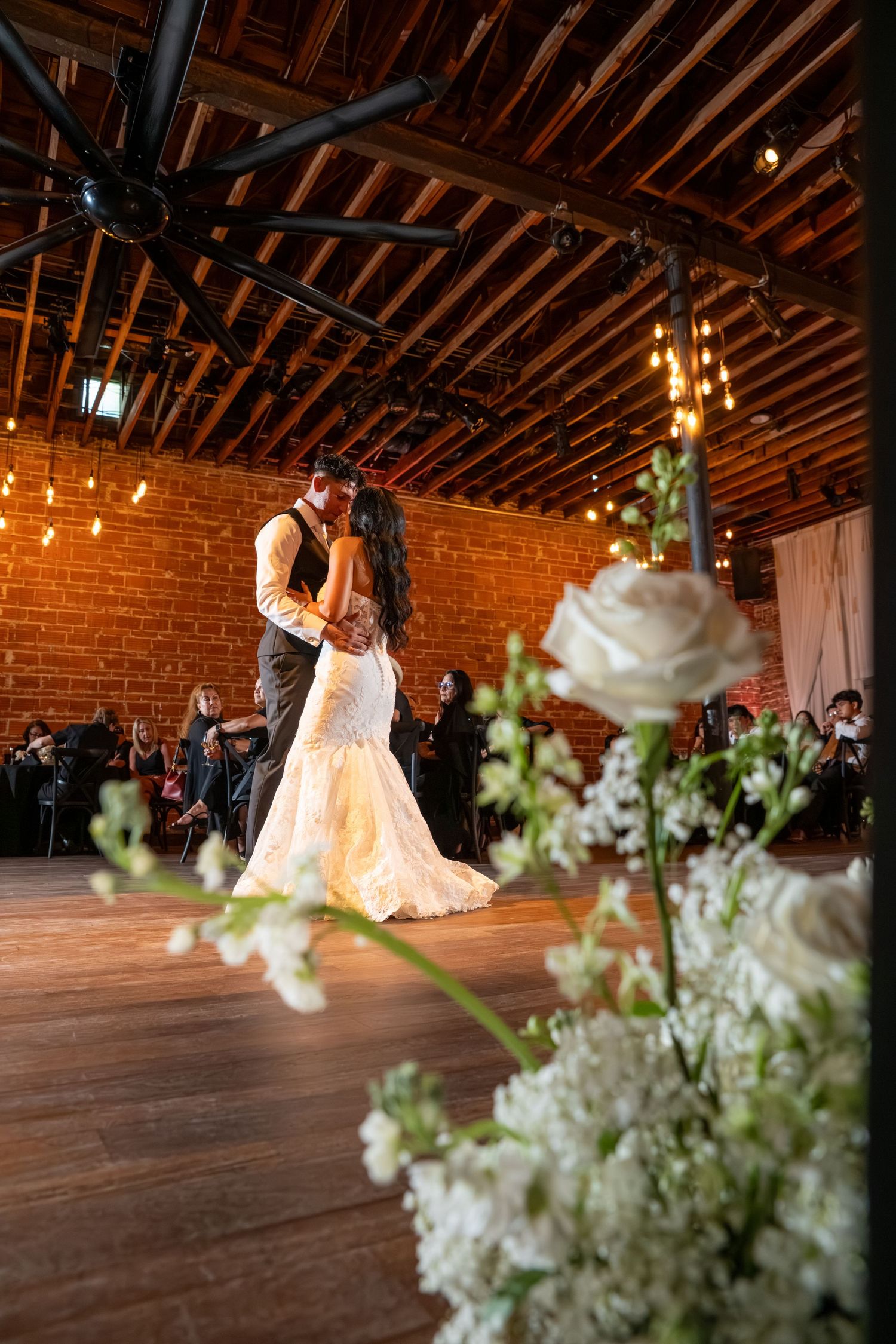 Bride and groom share first dance in rustic brick venue with warm Edison lights and white roses in foreground.