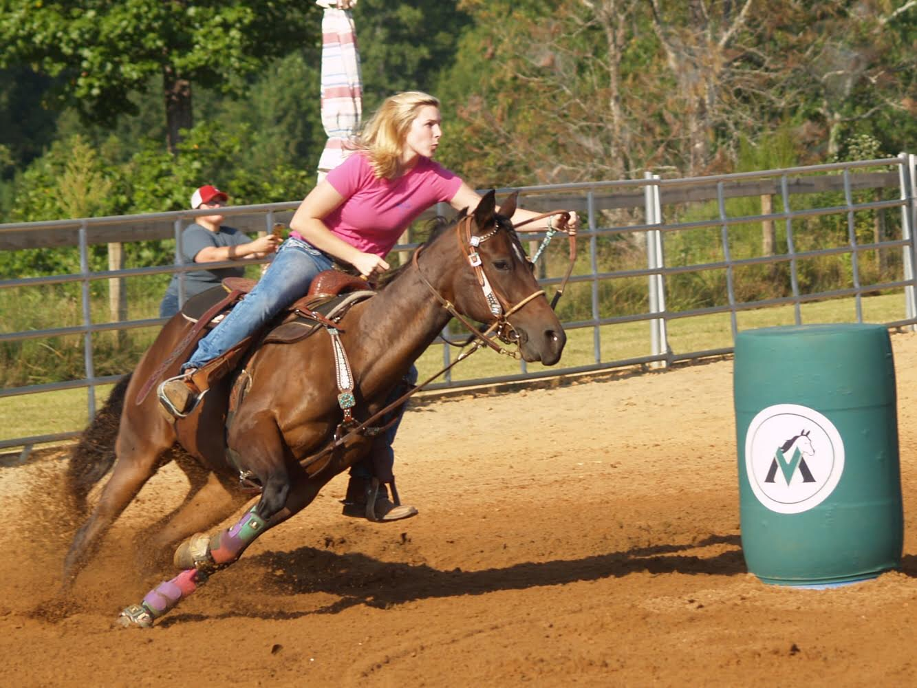 Woman in pink shirt barrel racing on a chestnut horse at an outdoor rodeo arena competition.