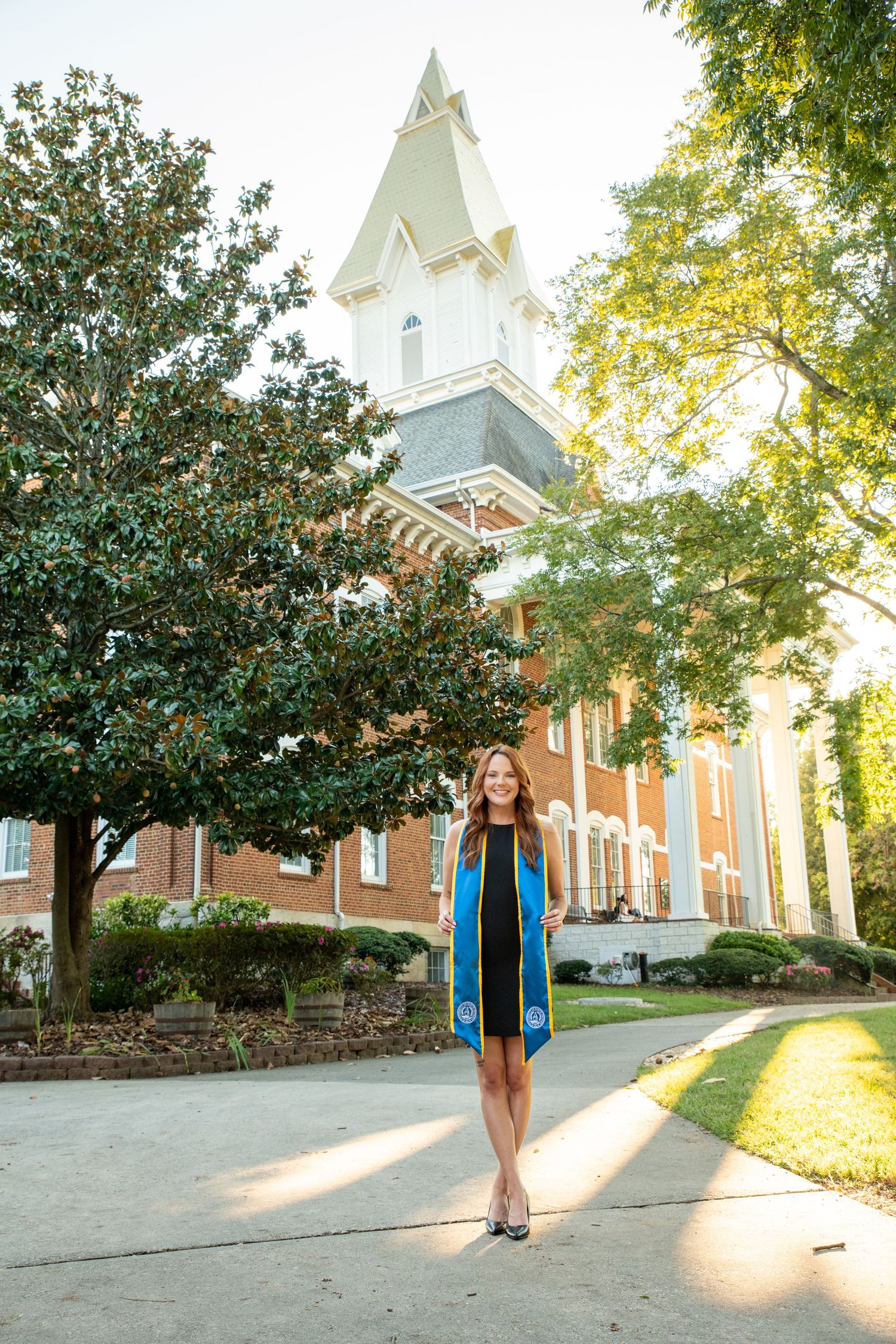 A graduate poses in a blue stole before a historic brick building with a white steeple on a sunny day.