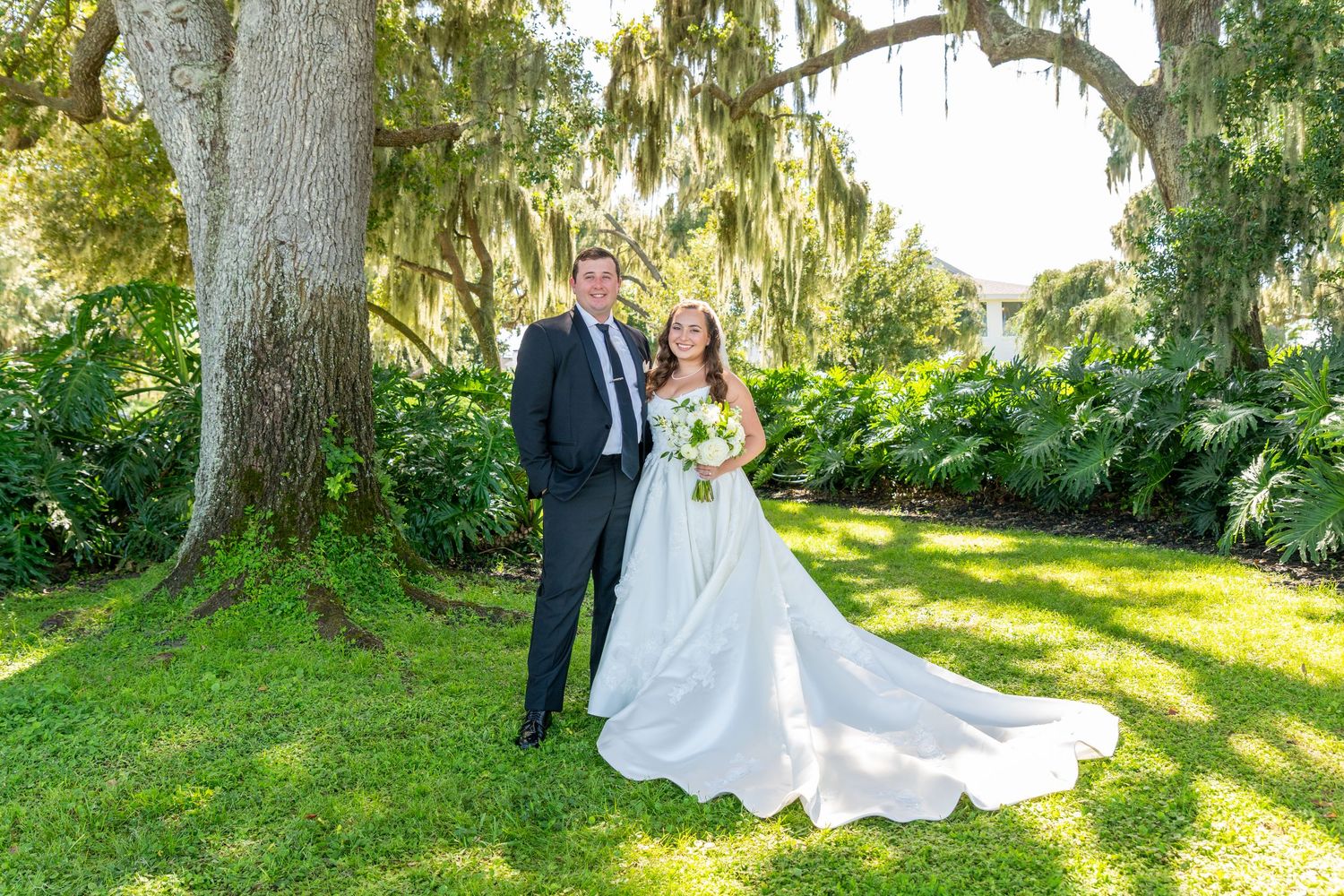 Bride and groom pose together under a moss-draped oak tree on lush green lawn in an outdoor wedding portrait.