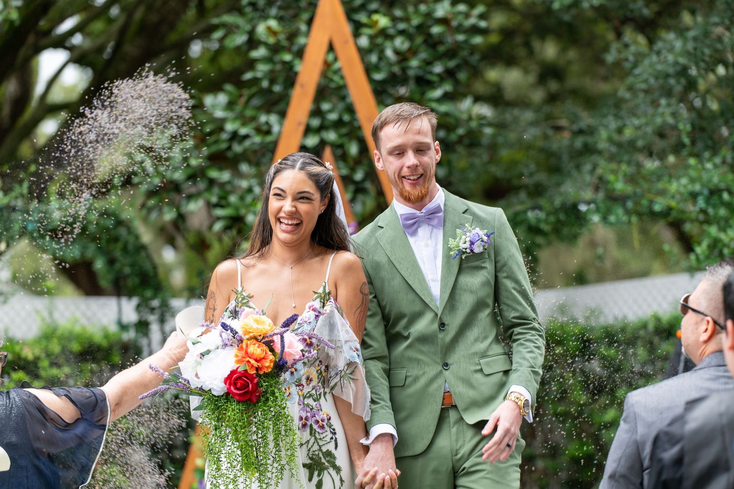 Happy newlyweds walk down the aisle at an outdoor garden wedding ceremony with colorful bouquet.