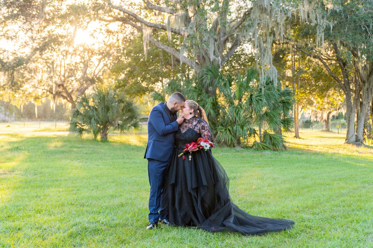 Couple shares romantic kiss in lush green park, bride in black gown with red bouquet, groom in navy suit, golden sunset.