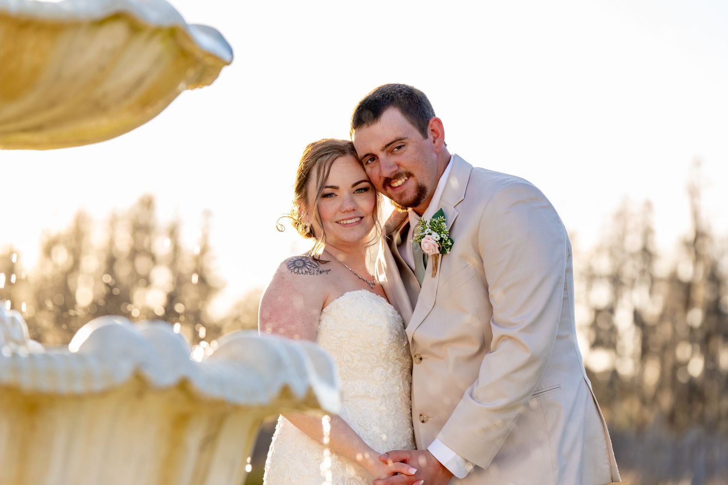 A smiling bride and groom pose together outdoors in golden sunlight on their wedding day.