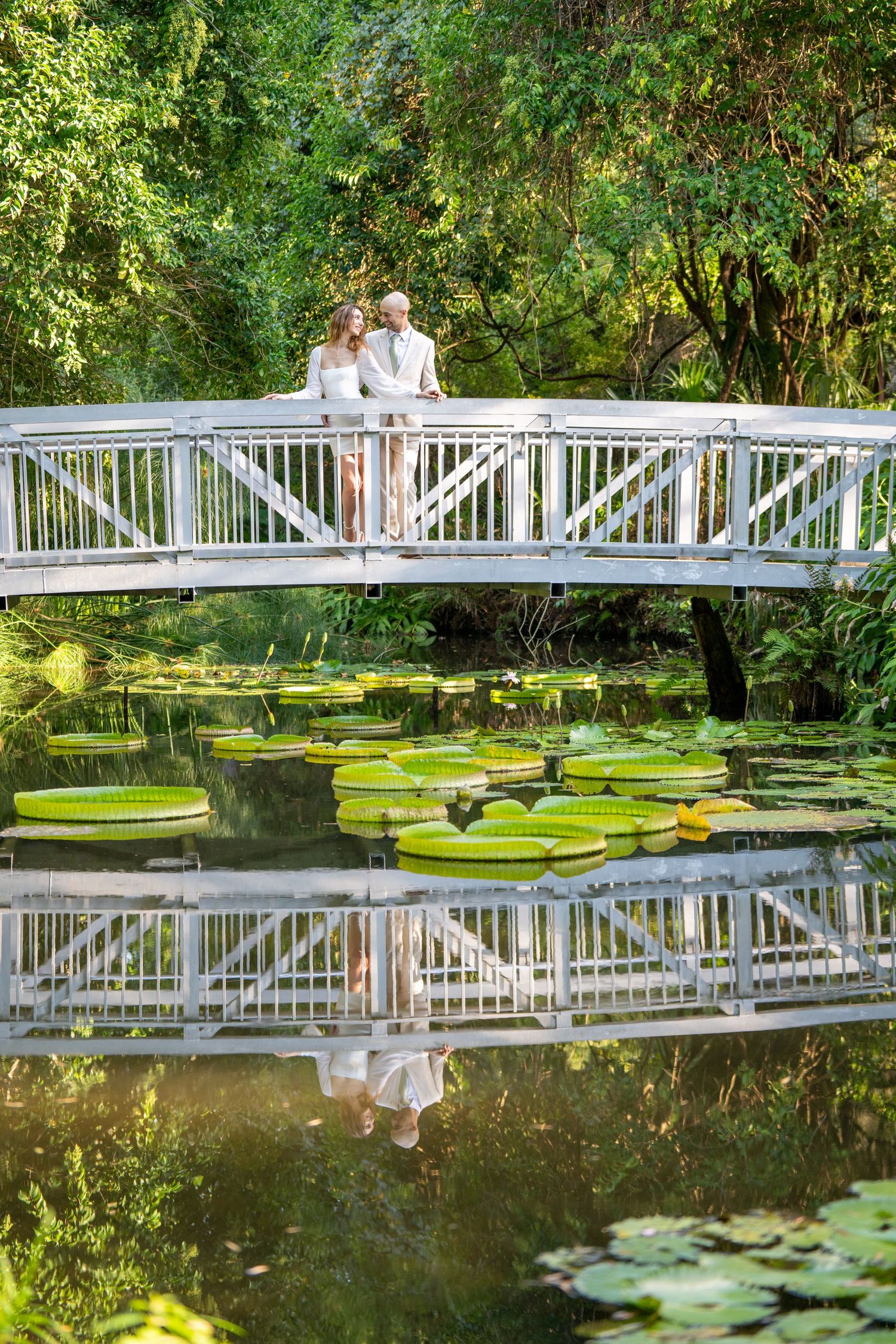 Couple standing on a white bridge reflected in a pond with large lily pads surrounded by greenery.