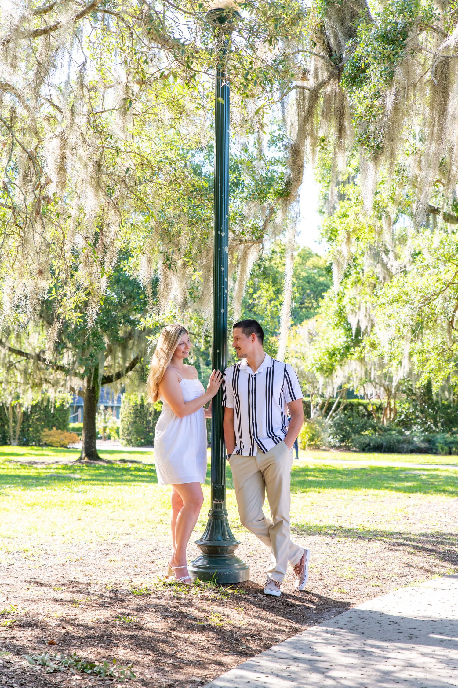 Couple standing by a green lamppost under Spanish moss-draped trees in a sunny park.