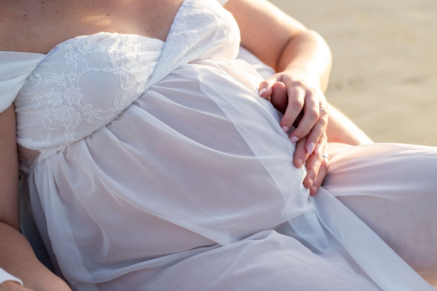 Pregnant woman in elegant white strapless maternity gown sitting gracefully on a beach.