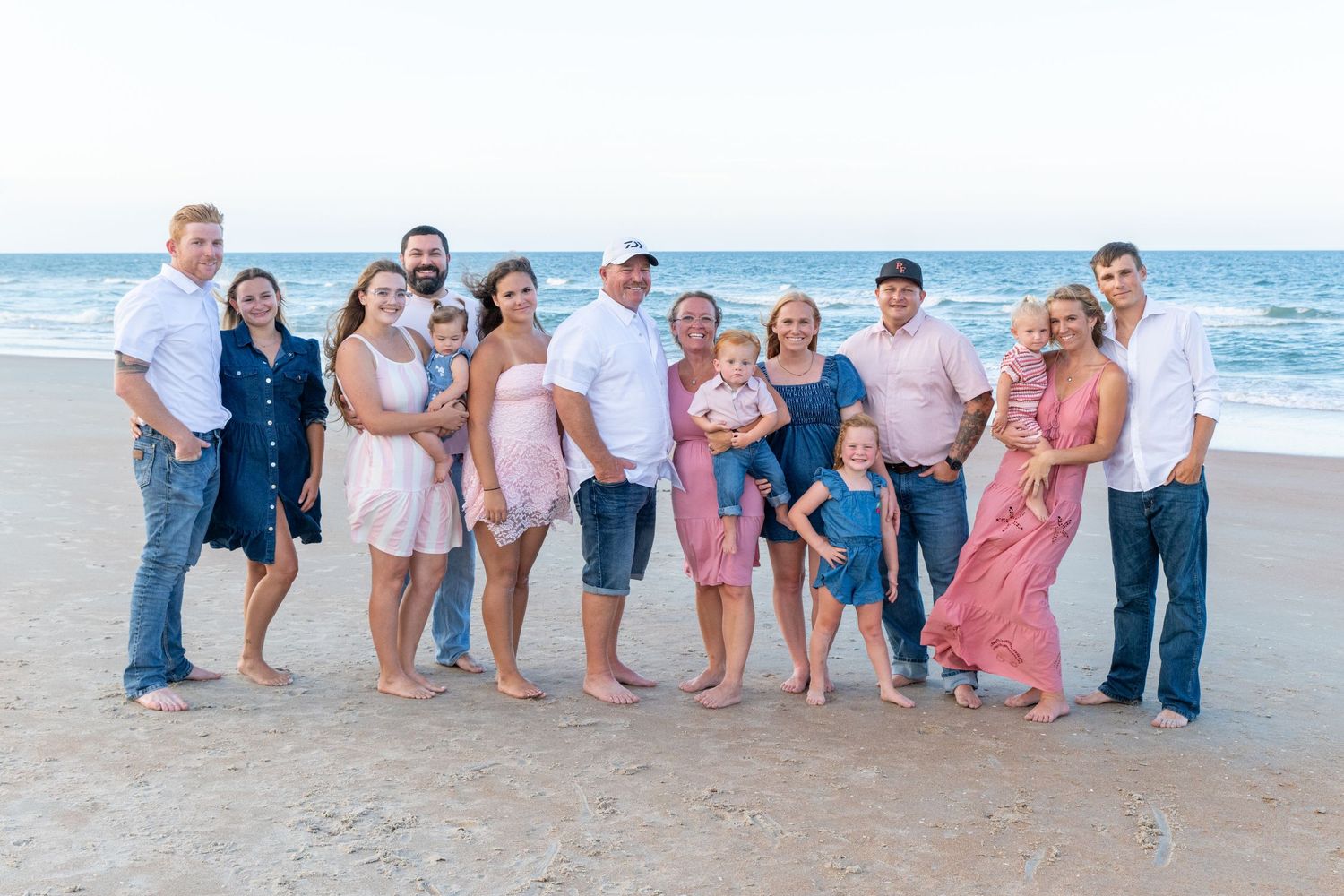 Large family group posing together on a sandy beach, smiling, wearing casual summer outfits in pink and blue tones.