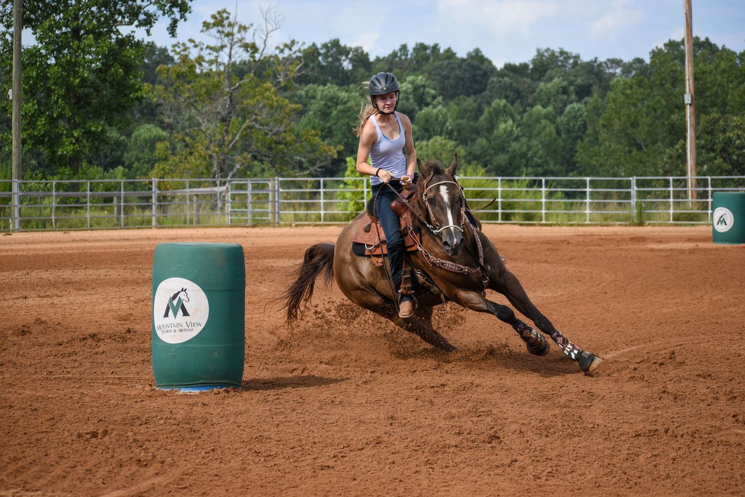 Young rider on a brown horse rounds a green barrel during a barrel racing competition in an outdoor dirt arena.
