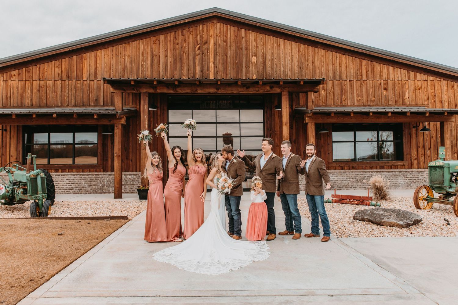 Wedding party posing in front of a rustic wooden barn venue, bridesmaids in coral gowns with groomsmen in brown suits.