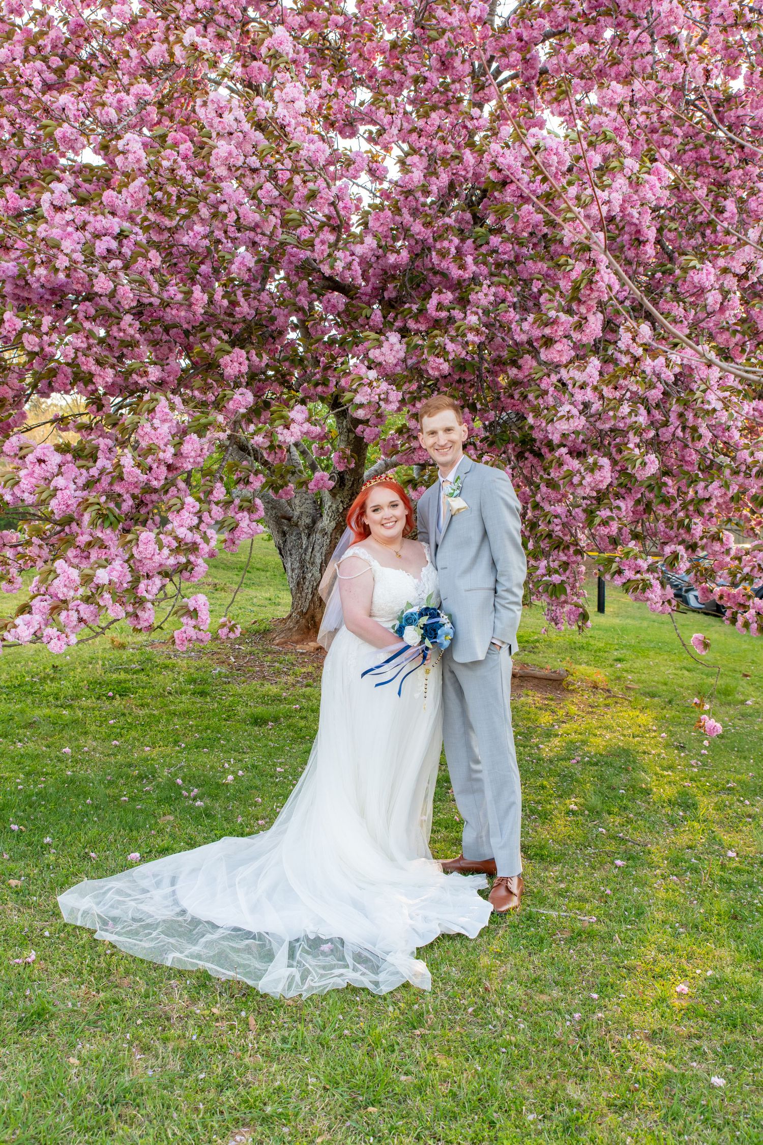 Bride and groom pose under a blooming pink cherry blossom tree on green grass during spring wedding.