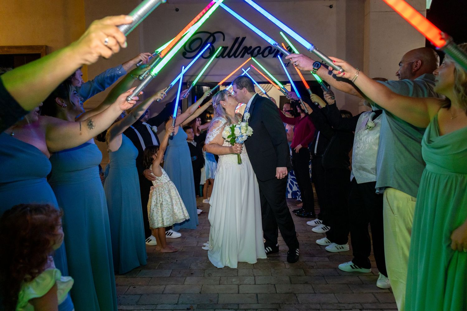 Bride and groom walk through glowing lightsaber tunnel held by wedding guests at elegant ballroom reception.