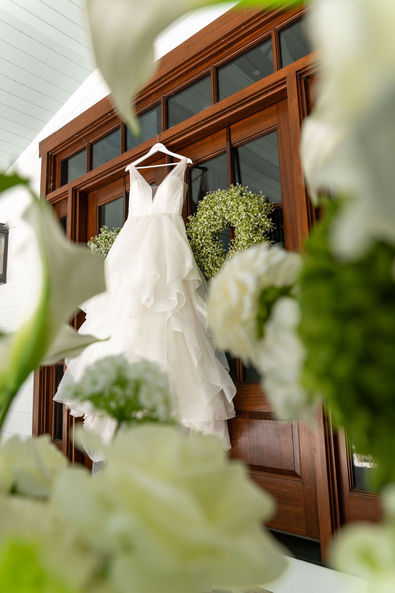 White wedding dress hanging on wooden door frame, framed by lush green wreath and white floral arrangements.