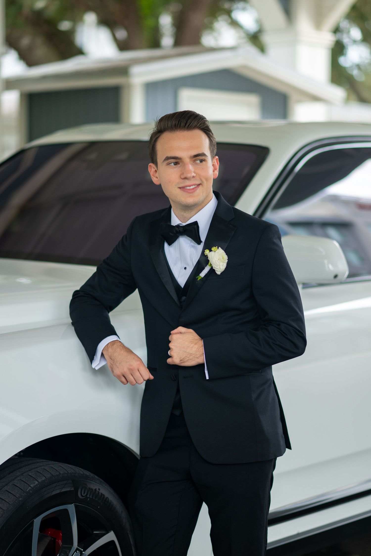 Groom in black tuxedo with white boutonniere leaning against white luxury car outdoors.