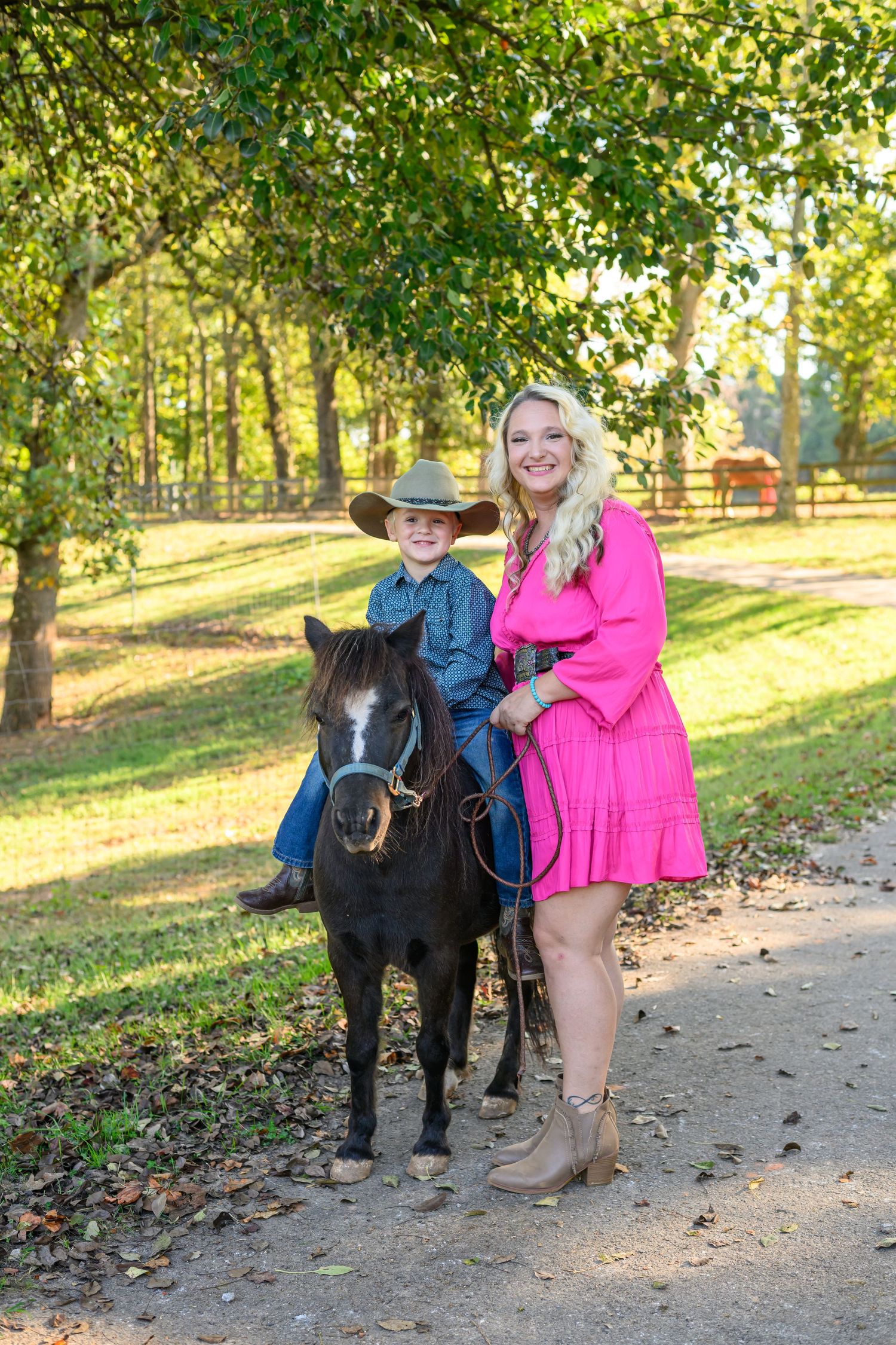 A woman in a pink dress and a boy in a cowboy hat pose with a small black pony on a sunny farm path.