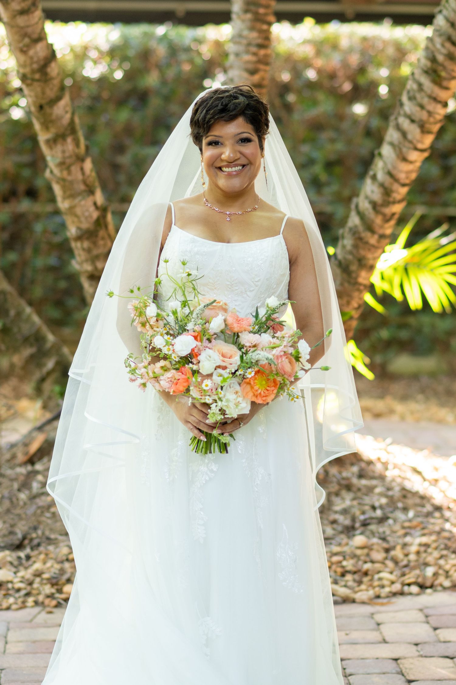 Smiling bride in white gown and veil holds peach and white floral bouquet outdoors among palm trees.