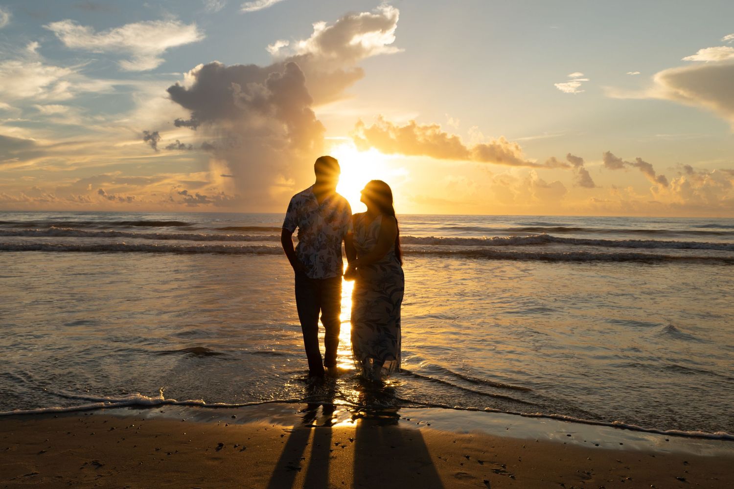 Couple holding hands on a sandy beach at golden sunset with dramatic clouds and ocean waves in the background.