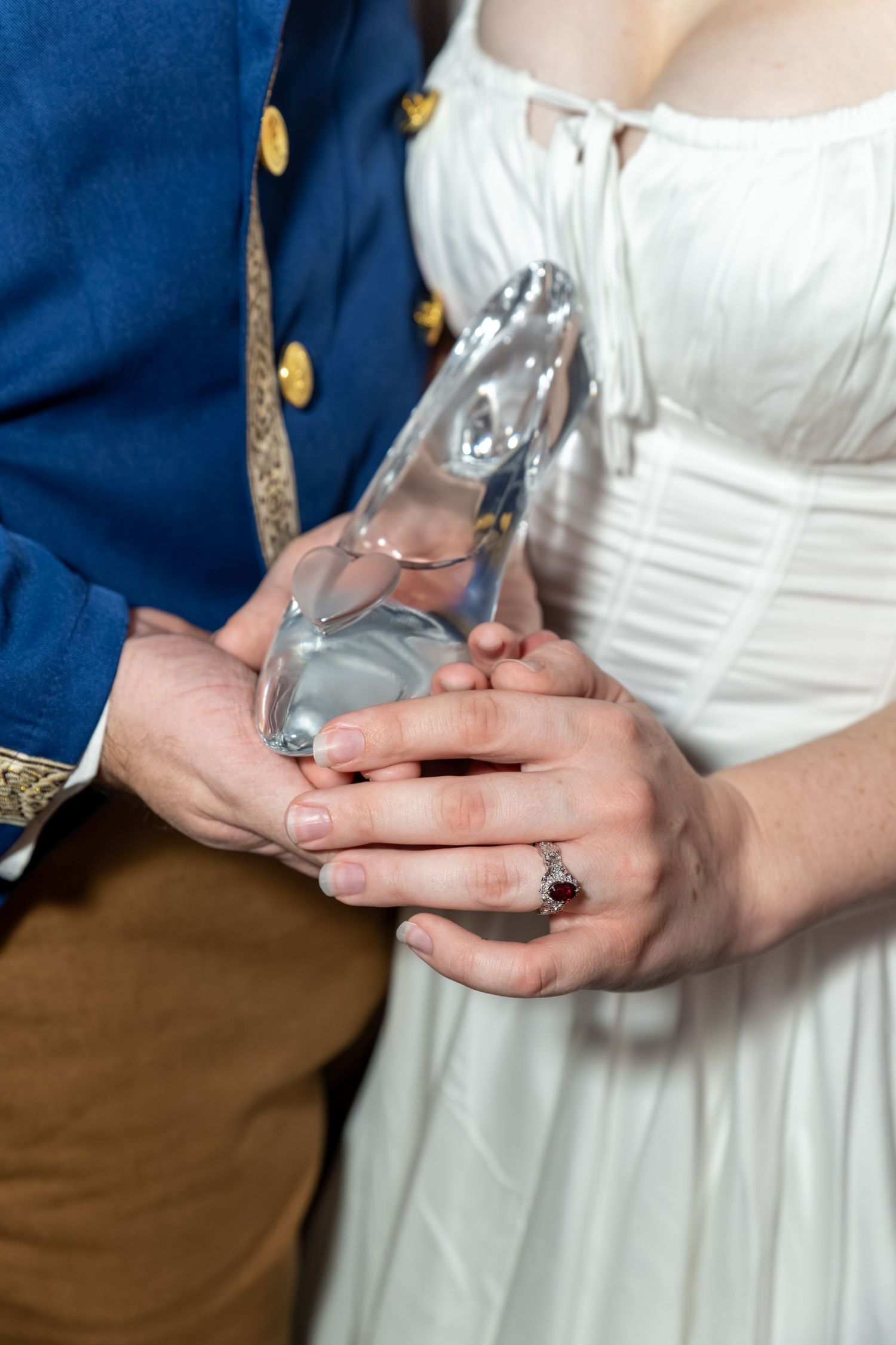 Couple in costume holding a glass slipper, woman wearing a red gemstone ring, Cinderella themed wedding photo.