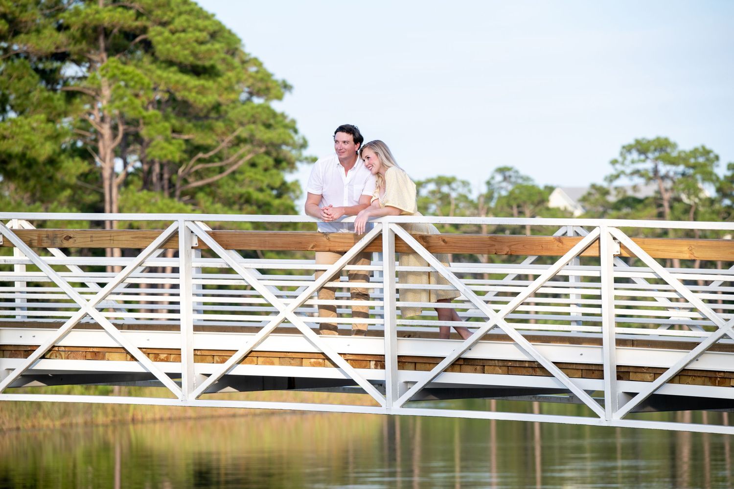 Couple leaning on white bridge railing over calm water surrounded by lush green trees.