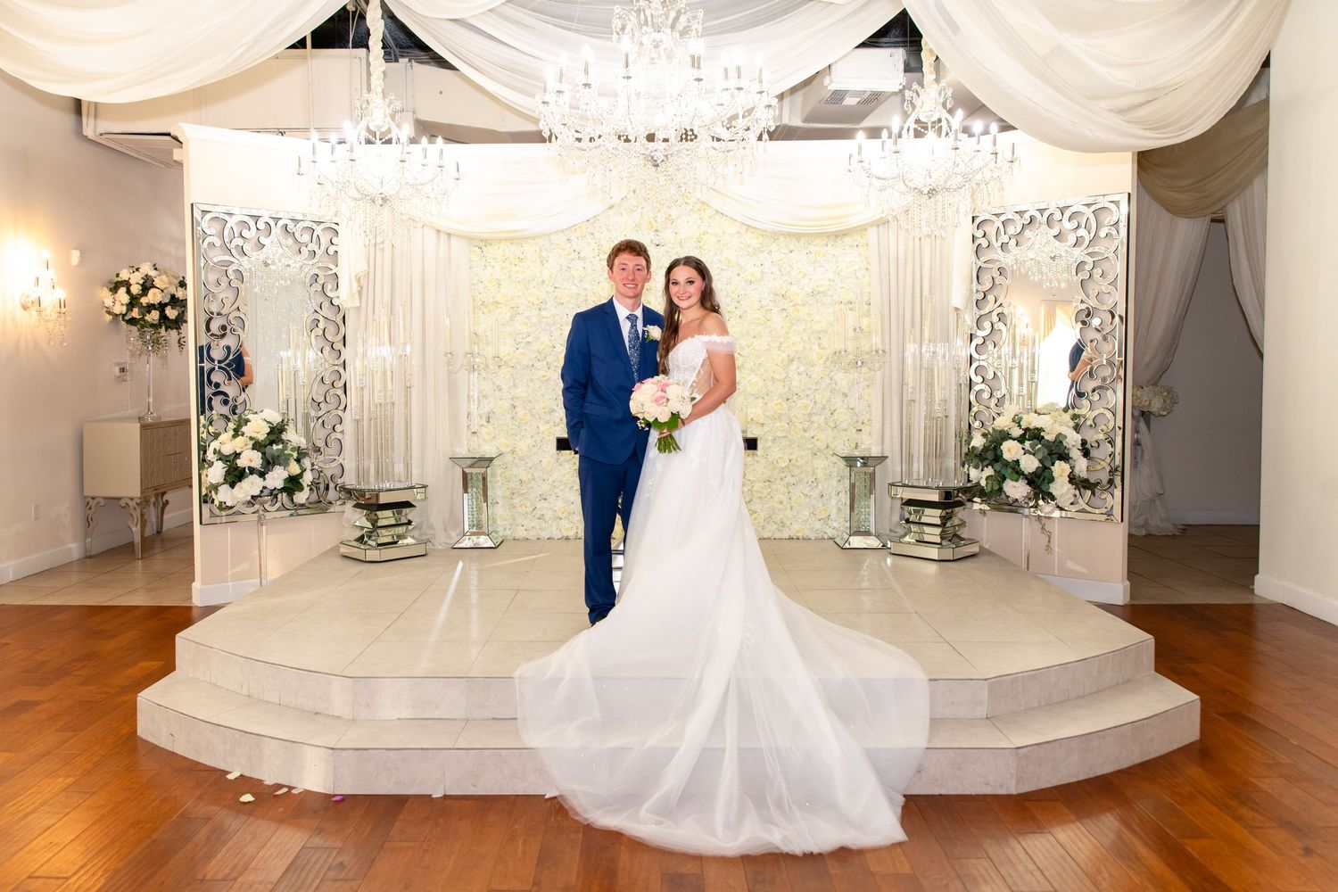 Bride and groom standing together at elegant white wedding altar with chandelier and floral decorations.