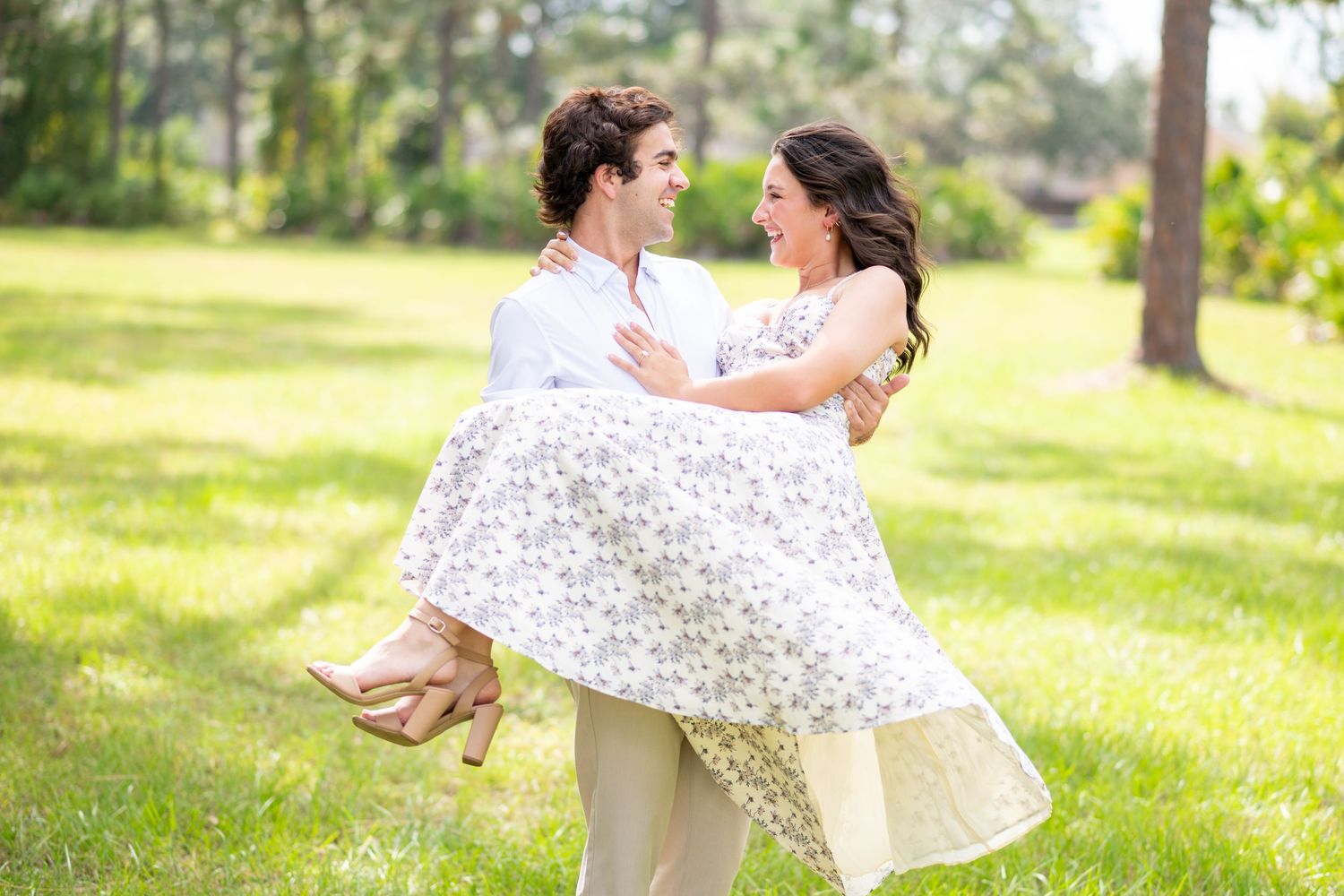 A man in white shirt lifts a woman in floral dress outdoors on lush green grass, sharing a romantic moment.