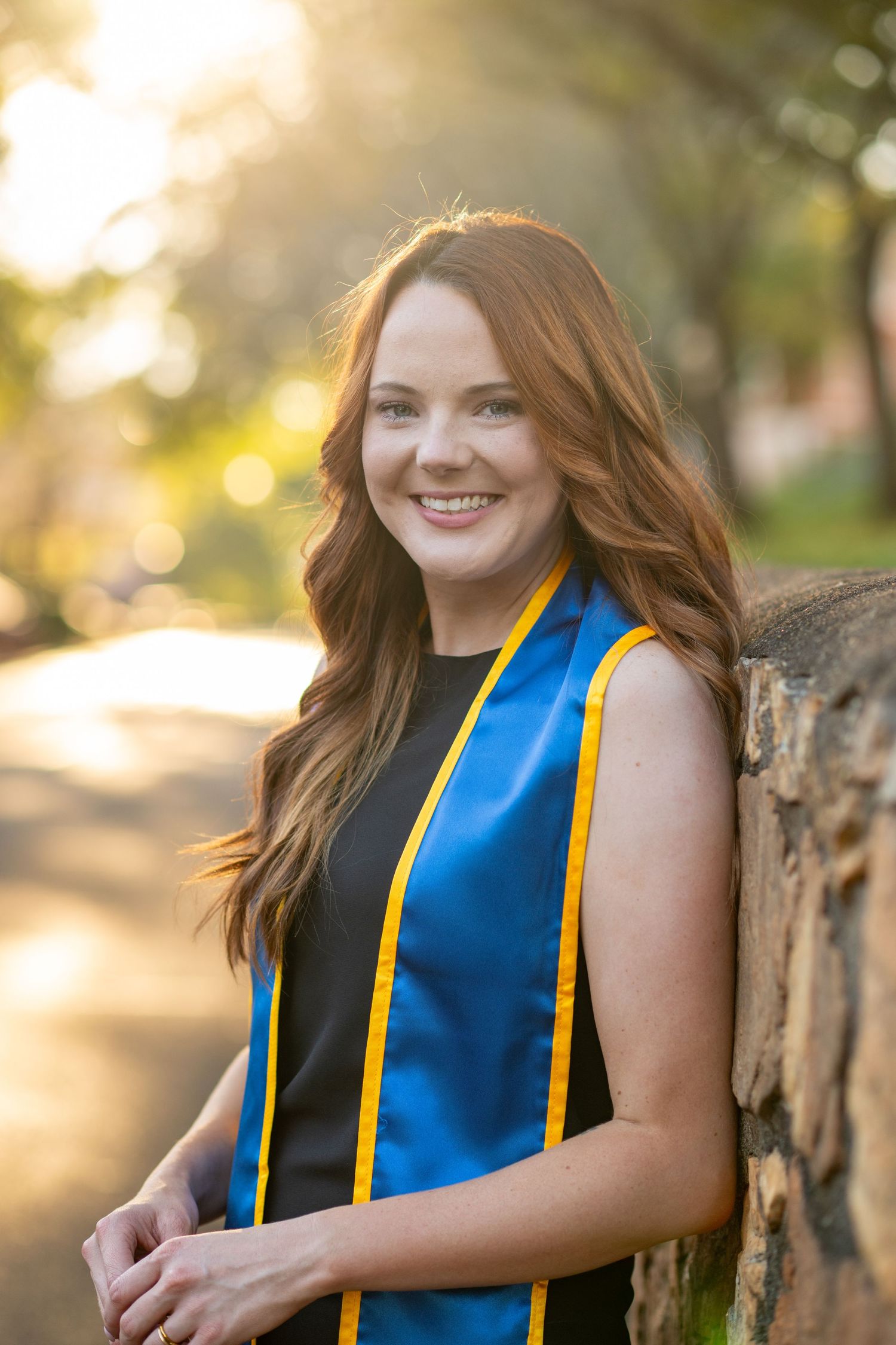 Young woman in blue and gold graduation stole smiling outdoors in warm sunlight, leaning against a stone wall.