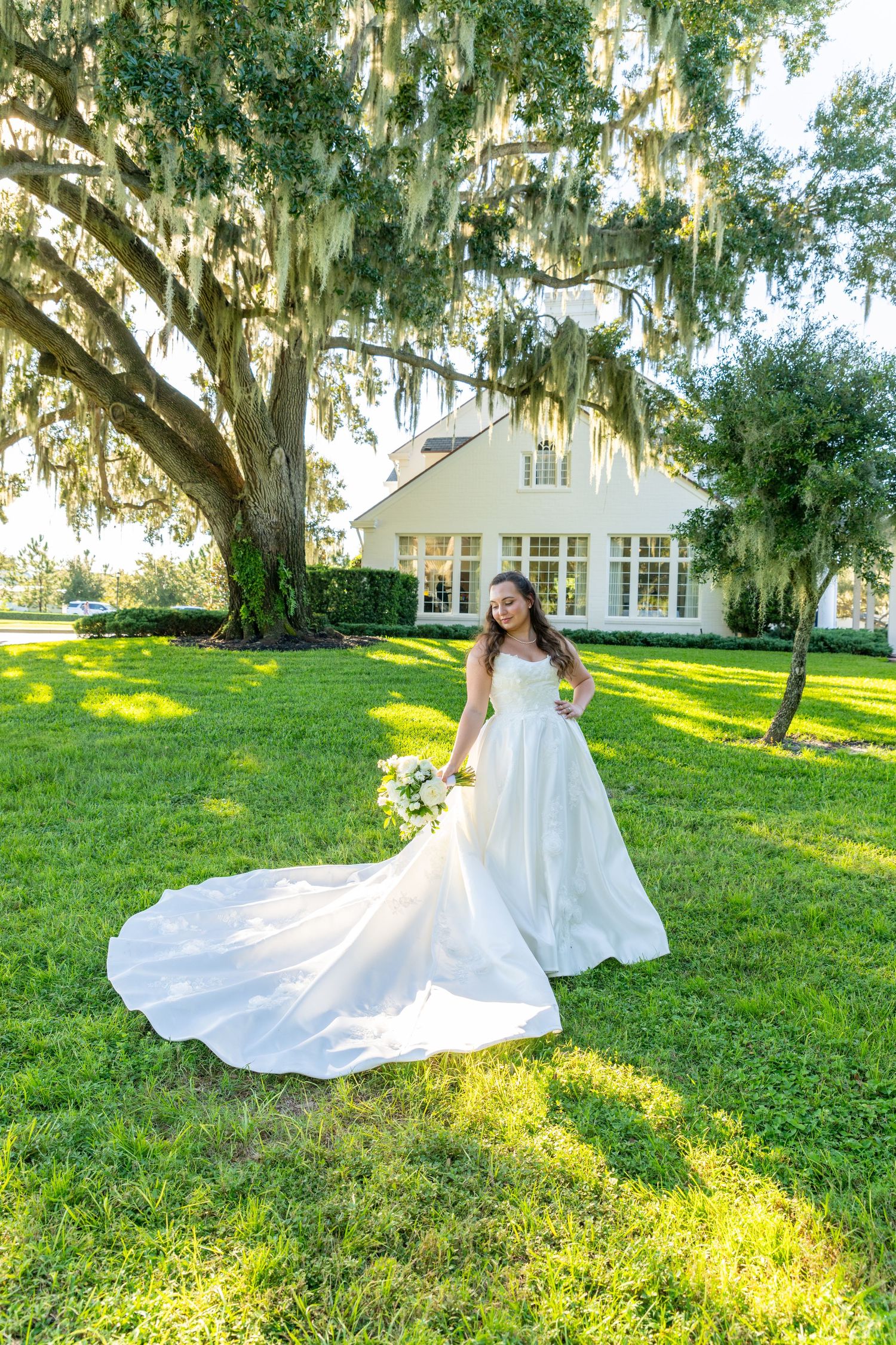 Bride in a white ball gown with long train stands on green lawn under Spanish moss oak tree near white venue.