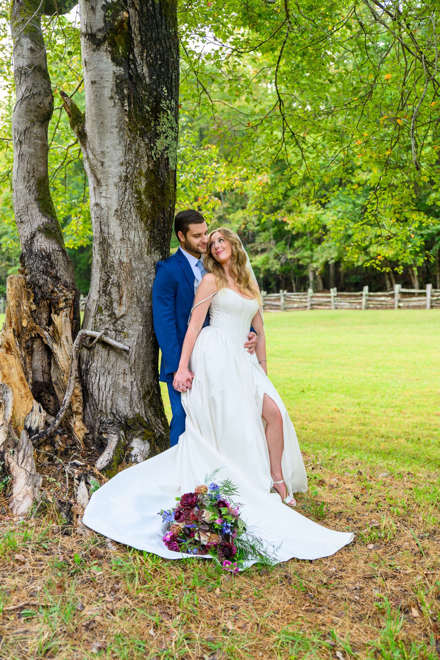 Bride in white gown and groom in blue suit pose romantically by a large tree in a lush green outdoor setting.