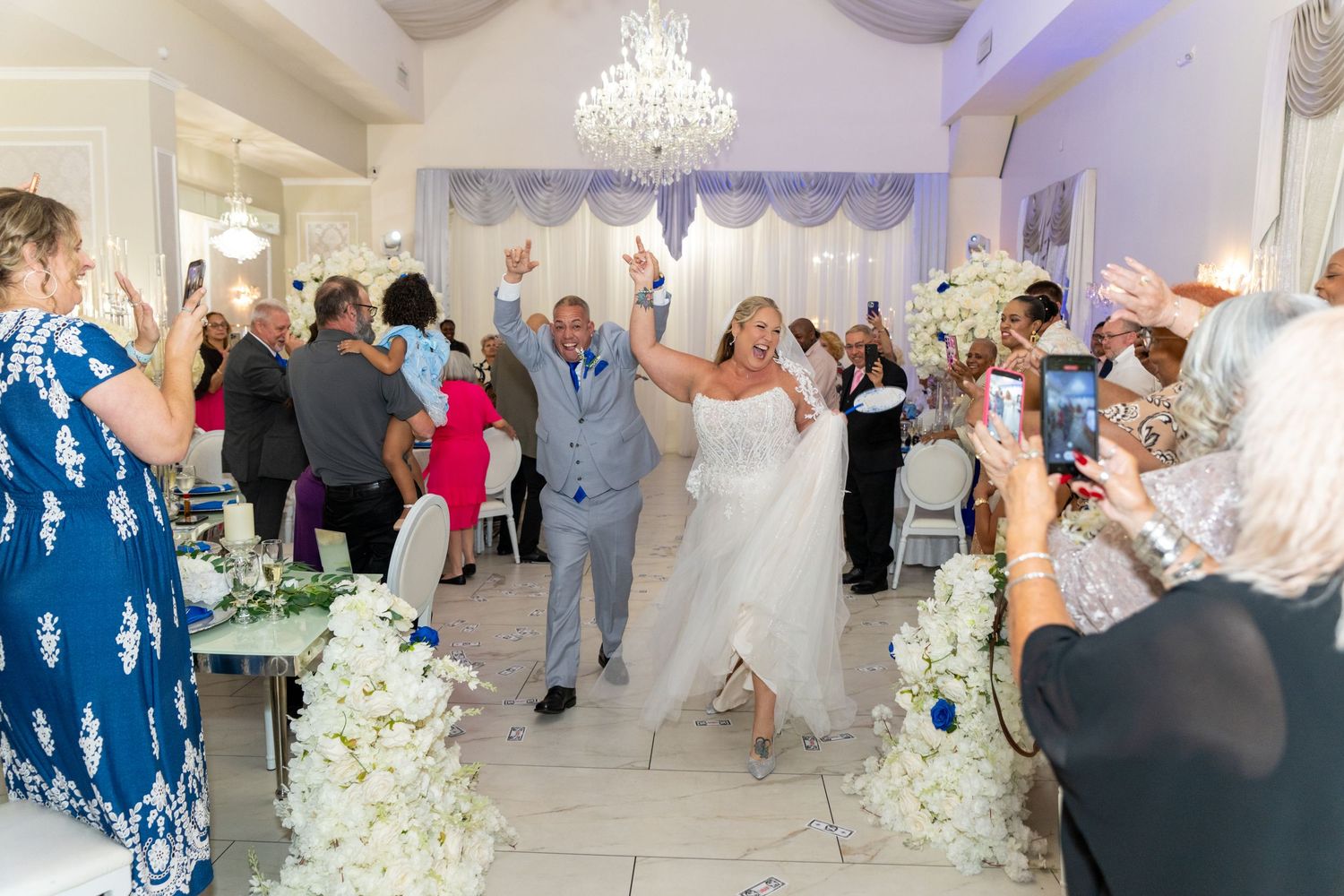 Bride and groom celebrate their wedding reception grand entrance with cheering guests in an elegant white floral venue.