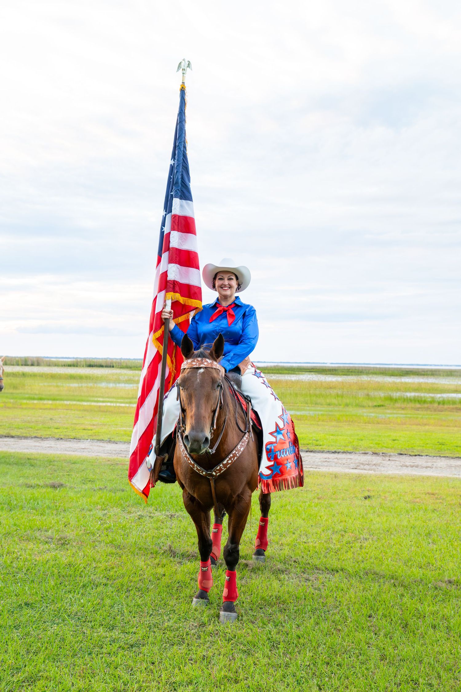 A smiling cowgirl in a blue shirt and white hat rides a horse while proudly carrying a large American flag outdoors.