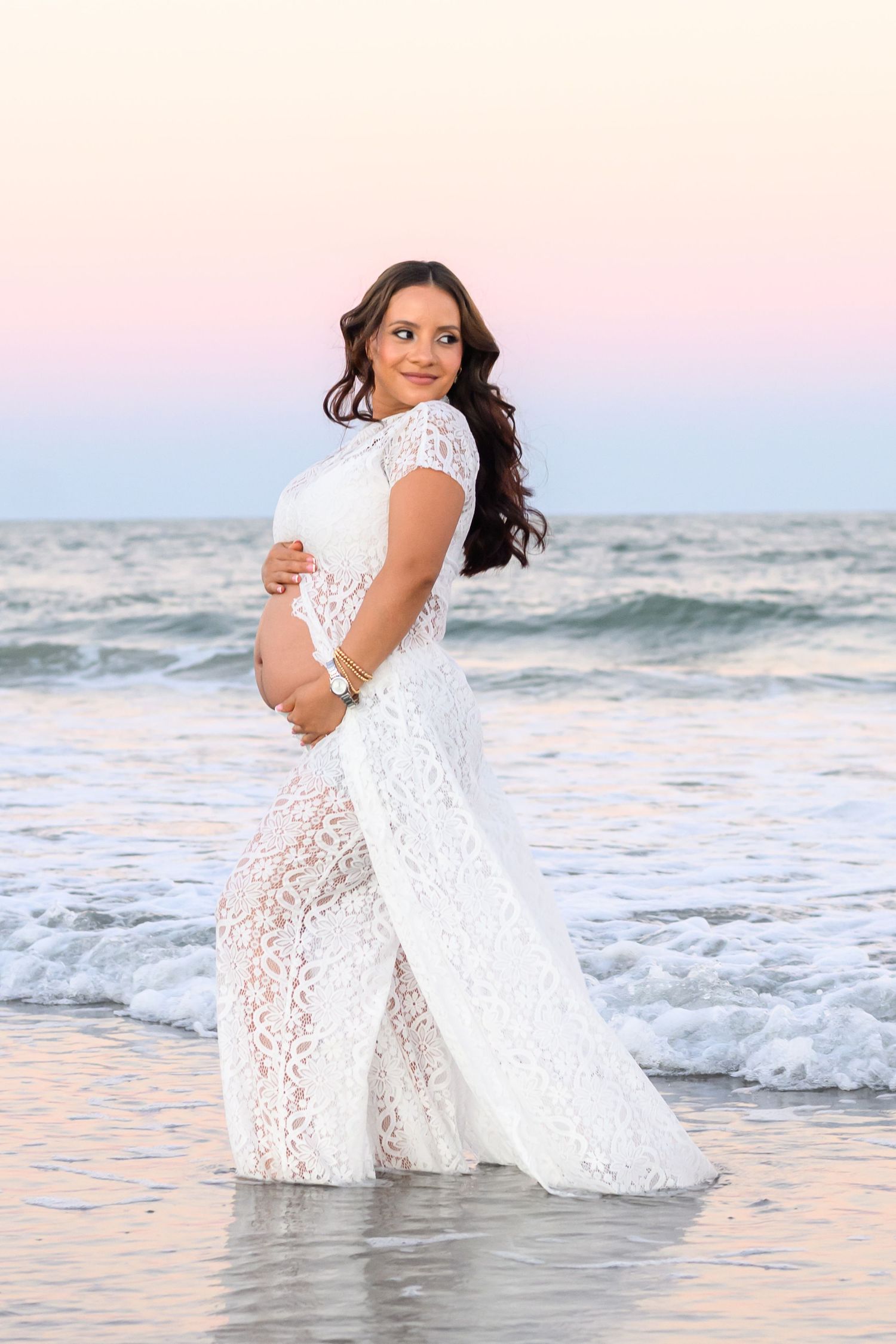 Pregnant woman in a white lace maternity gown cradling her belly while standing in shallow ocean waves at sunset.