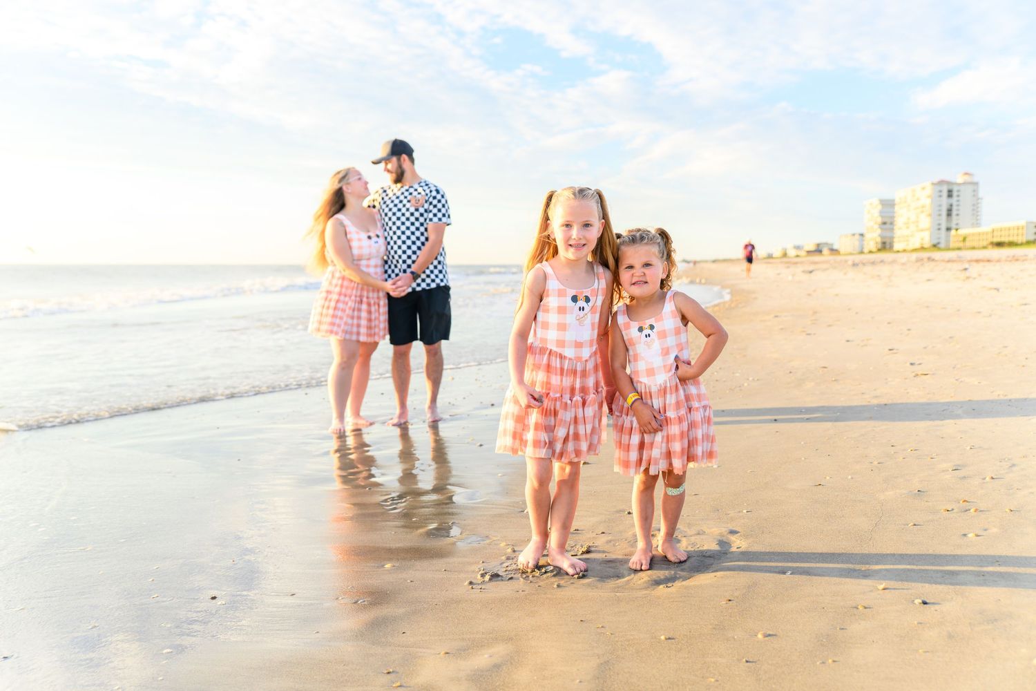 Two young girls in matching pink plaid outfits stand on a sunny beach while their parents embrace by the ocean in the background.