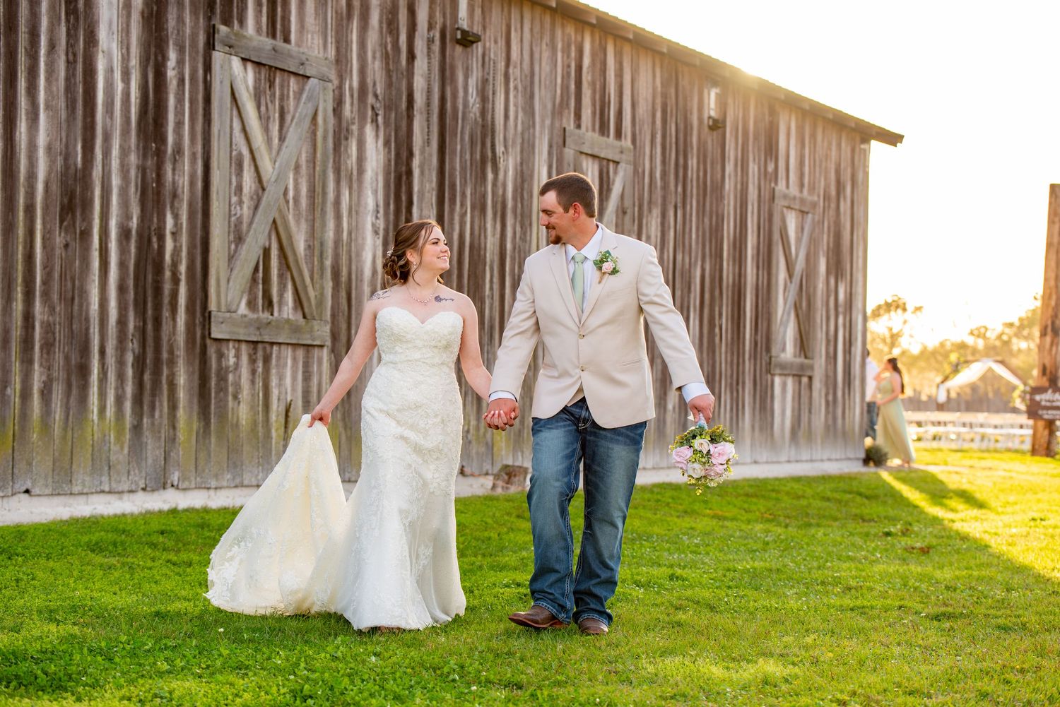 Bride and groom holding hands walking past rustic wooden barn on green grass at outdoor wedding.