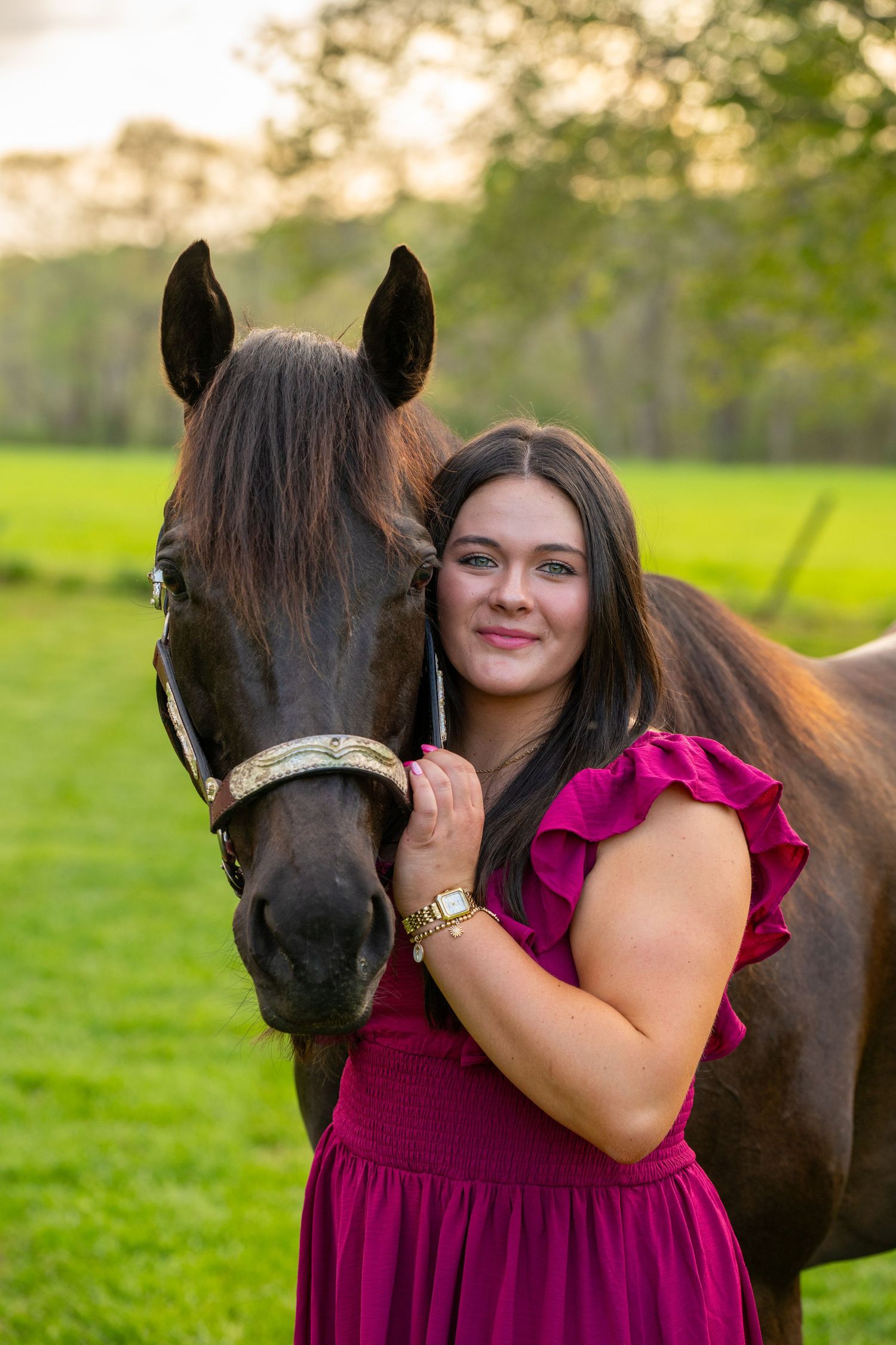 Young woman in pink dress embracing a dark horse in a lush green field at sunset.