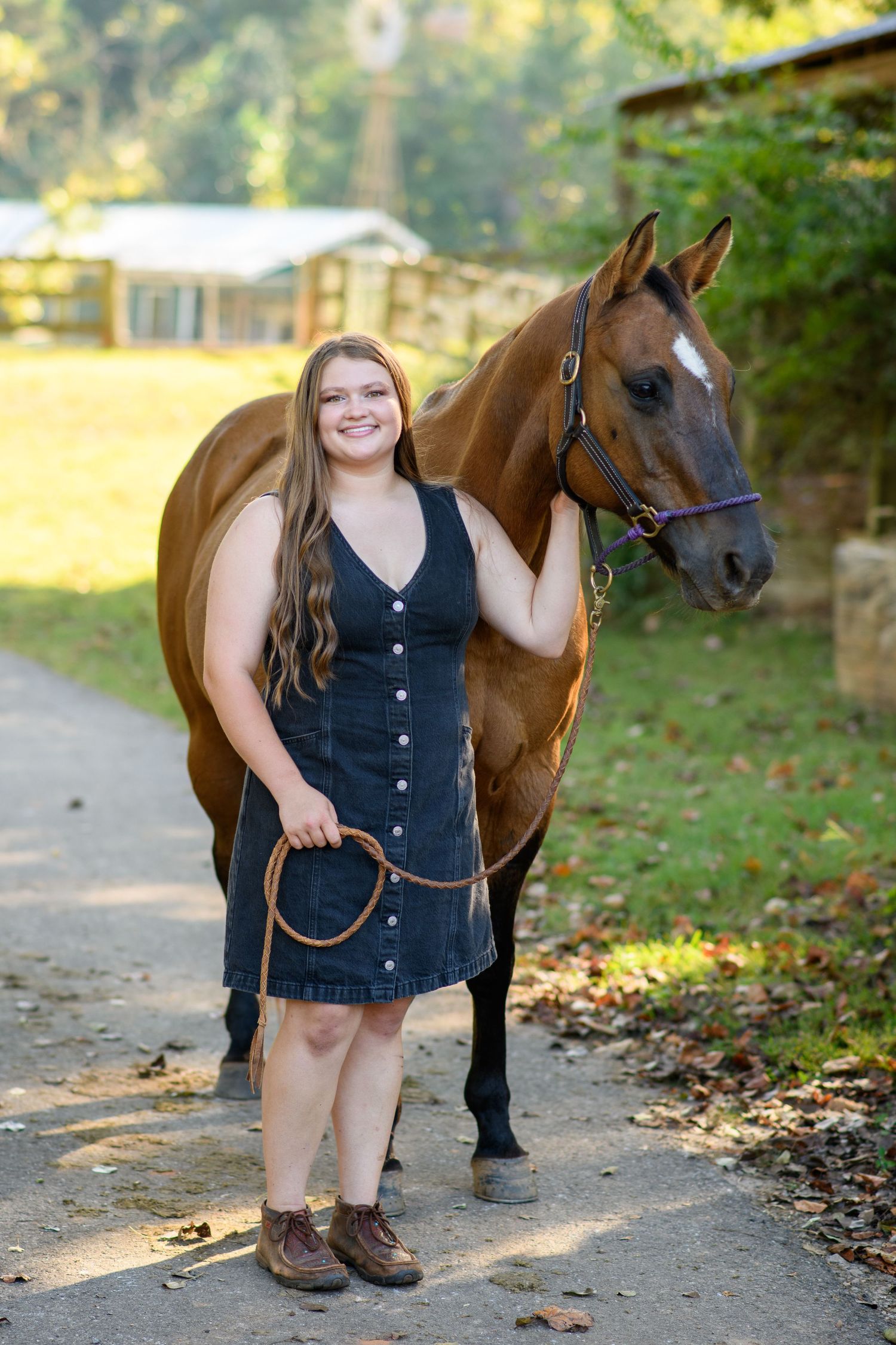 Young woman in denim dress holds lead rope beside chestnut horse on a tree-lined farm path.