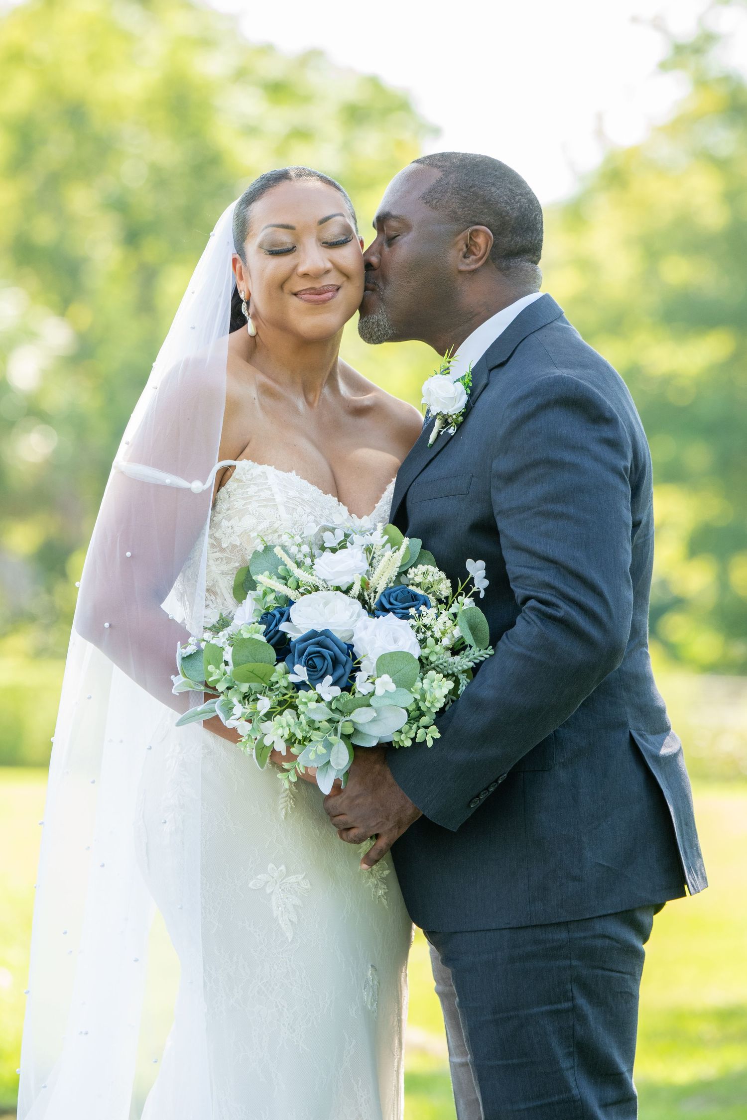 Groom in navy suit kisses bride in white lace gown holding blue and white bouquet outdoors on wedding day.