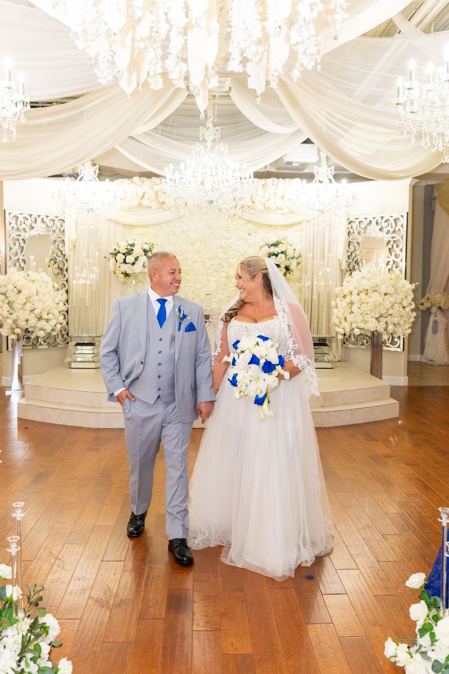 Bride and groom walking hand in hand in elegant white wedding venue with crystal chandeliers and floral arrangements.