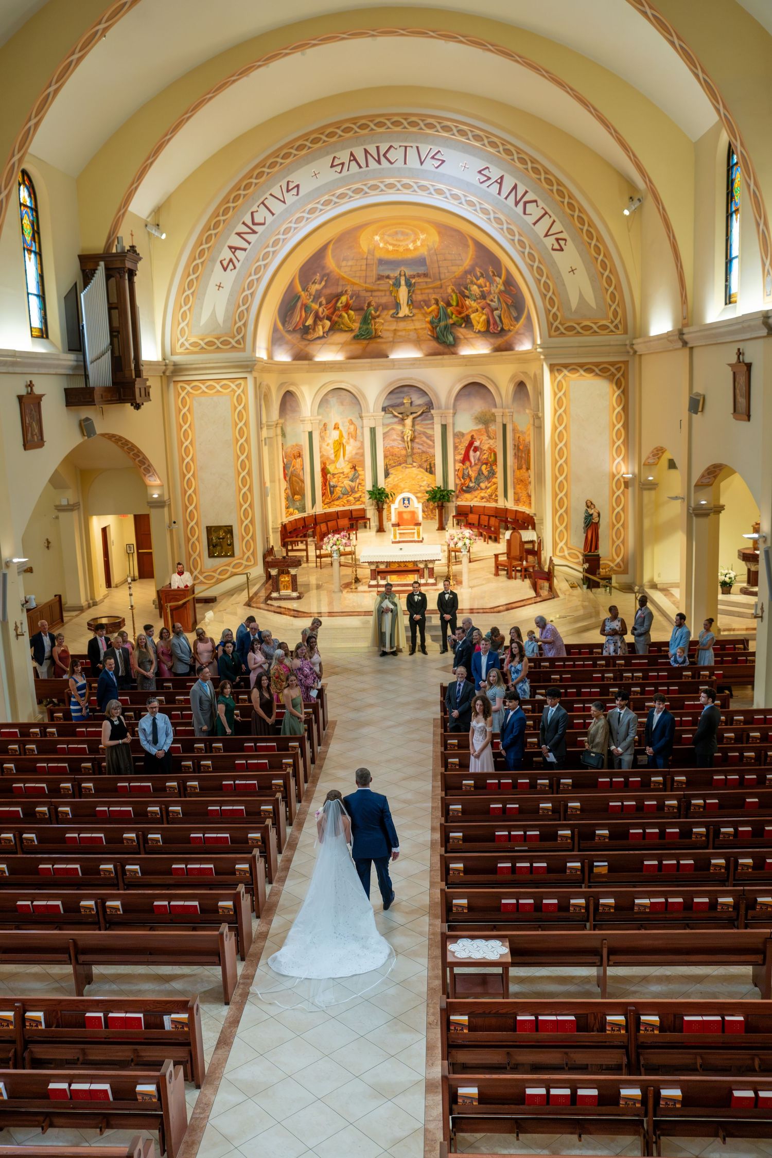 Bride and groom walk down the aisle of an ornate Catholic church with golden altar and Sanctus inscriptions.