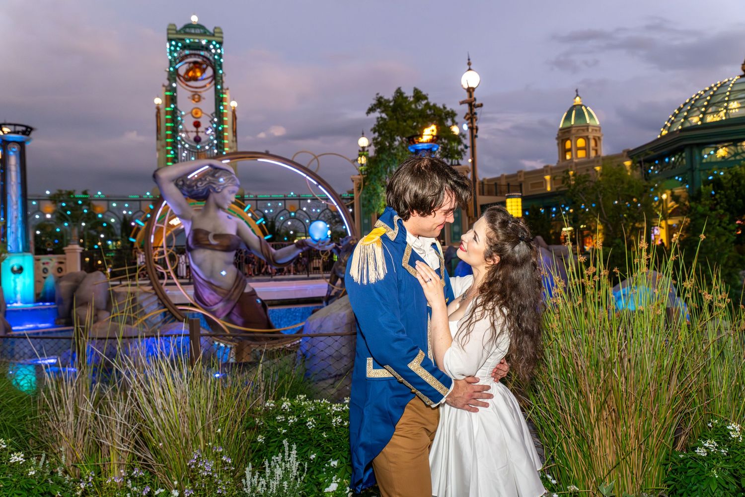 Couple in fairy tale costumes embrace near a mermaid fountain at a magical theme park at night.