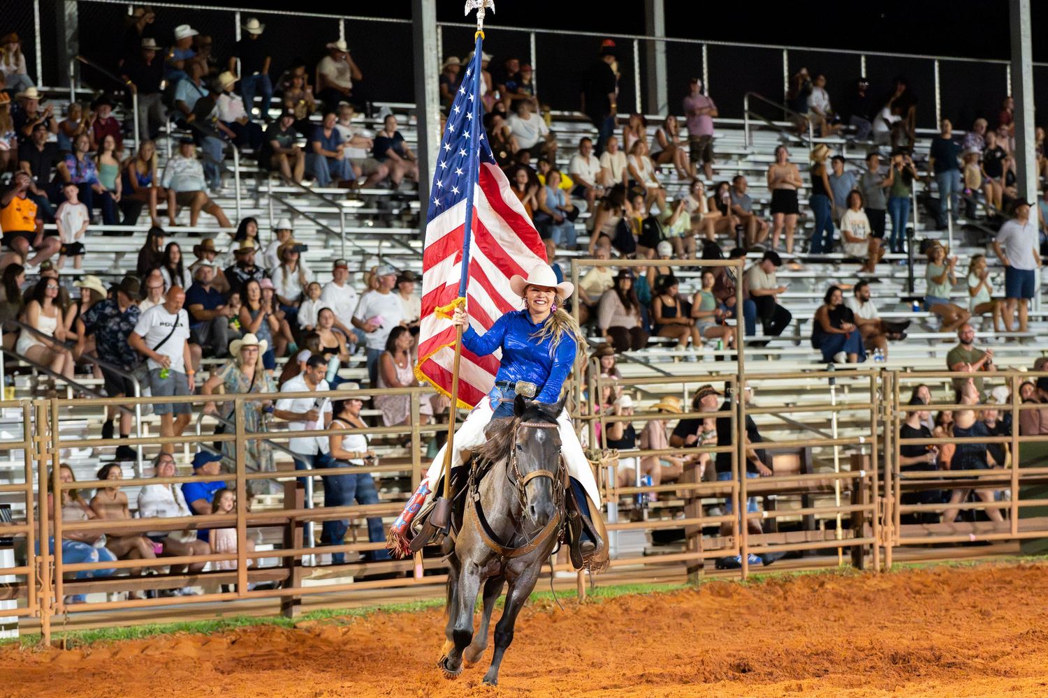 Rodeo performer on horseback carrying American flag before packed grandstands at night under bright arena lights.