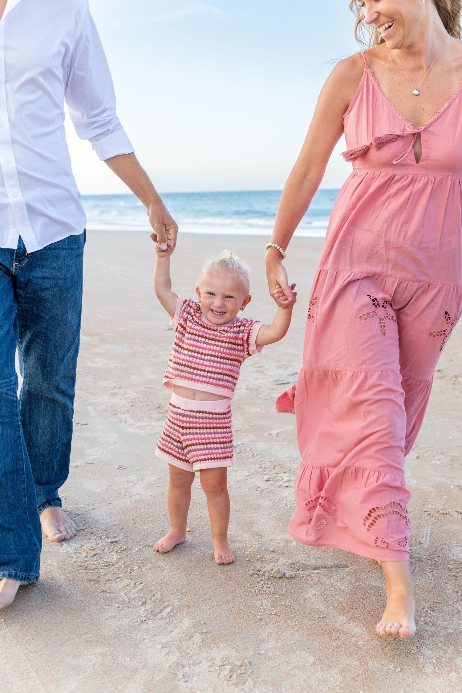A happy toddler holds parents hands on a sandy beach, wearing a pink outfit, with ocean waves in the background.
