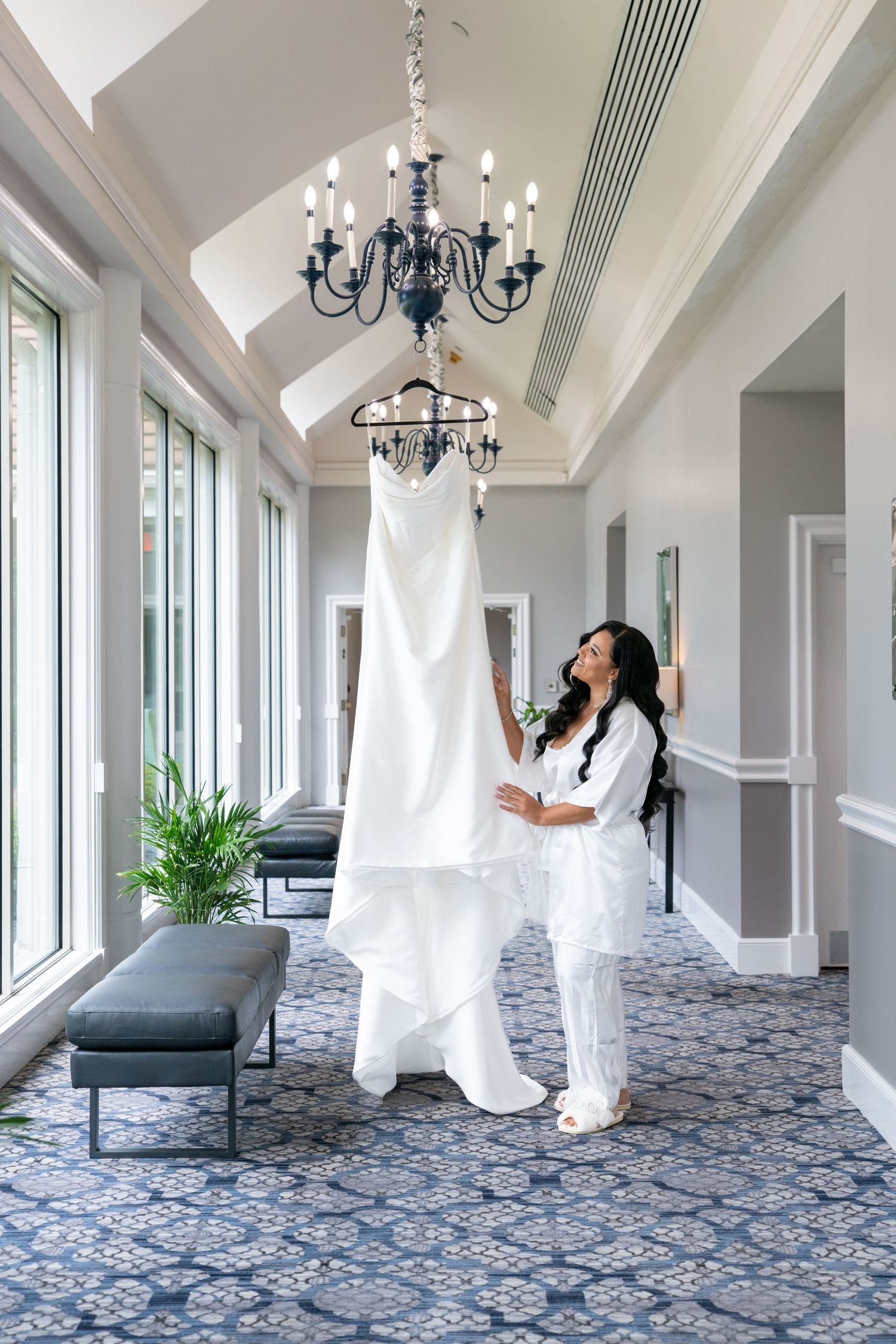 Bride in white robe admiring her wedding gown hanging from chandelier in elegant venue with blue patterned carpet.