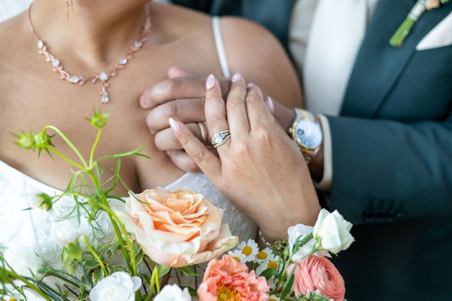 Newlywed couple holding hands showing wedding rings with a bridal bouquet of peach and white flowers in foreground.