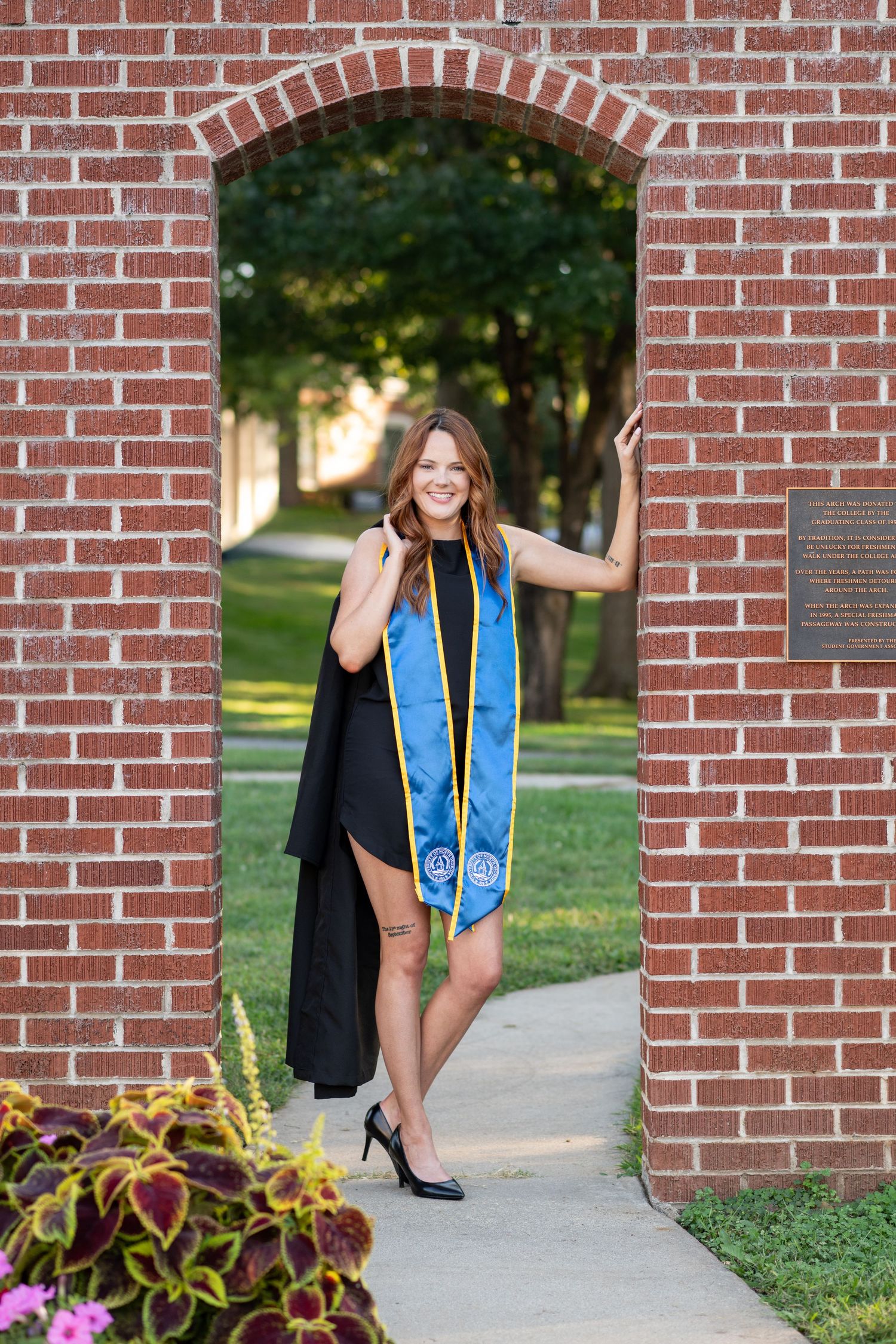 A smiling graduate poses in a brick archway wearing a black dress, blue graduation stole, and heels on a sunny day.